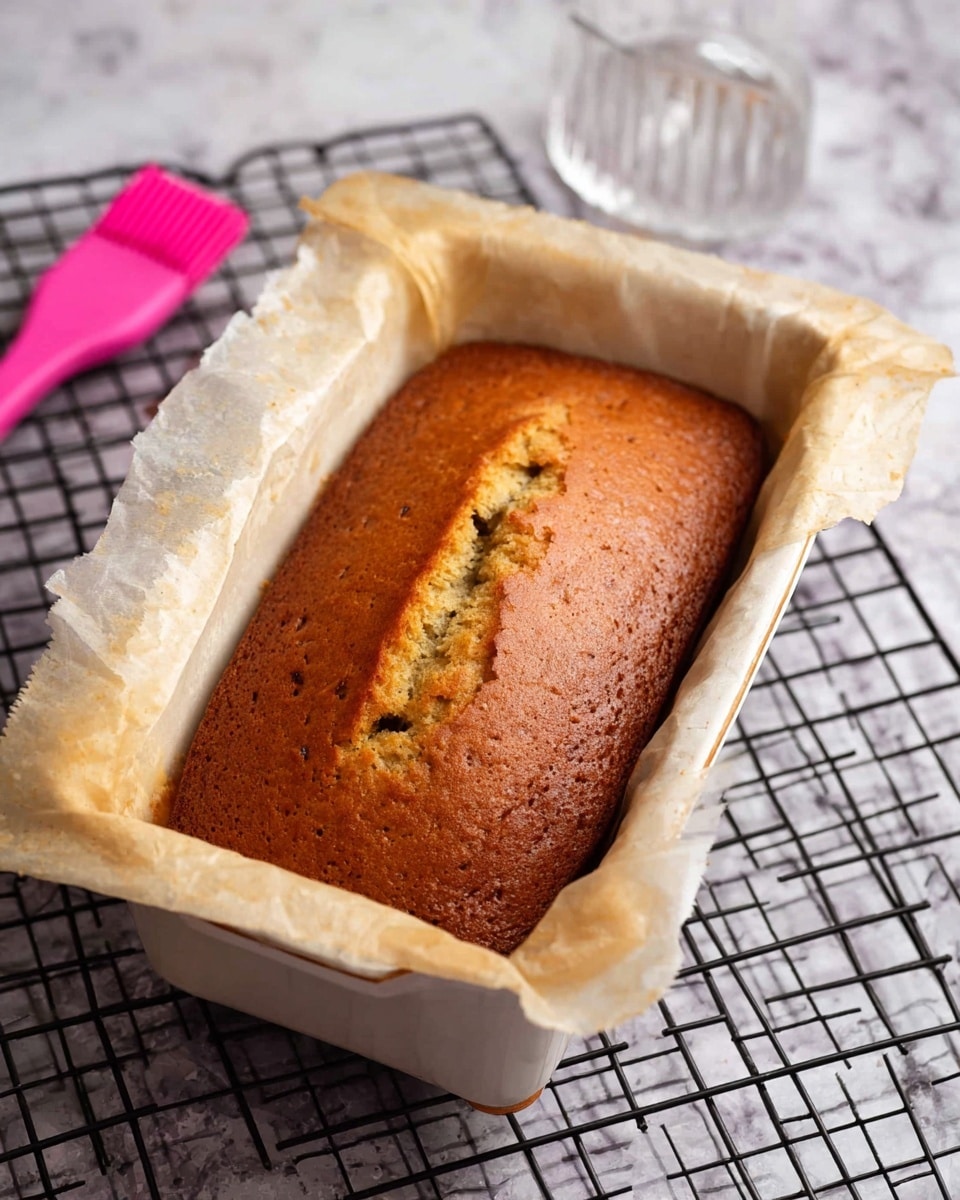 A single loaf of golden brown bread is shown in a white loaf pan lined with slightly browned parchment paper. The bread has a crack down the middle with small holes from air bubbles on the surface. The pan is resting on a black cooling rack over a white marbled surface. In the background, there is a pink silicone brush and a transparent measuring cup. Photo taken with an iphone --ar 4:5 --v 7
