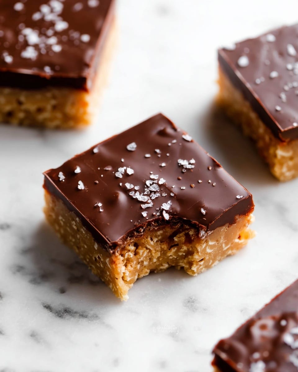 The image shows three square bars placed on a white marbled surface. Each bar has two layers: a bottom layer that is golden brown with a bumpy and textured look, resembling a crispy or crunchy base, and a top layer that is smooth and shiny dark chocolate with small textured spots and a few coarse white salt flakes sprinkled on top. One bar is in the foreground and has a bite taken out of its front left corner, exposing the thick golden brown base beneath the chocolate. The other two bars are partly visible, with one near the top edge and the other in the bottom left corner, both fully intact. Photo taken with an iphone --ar 4:5 --v 7