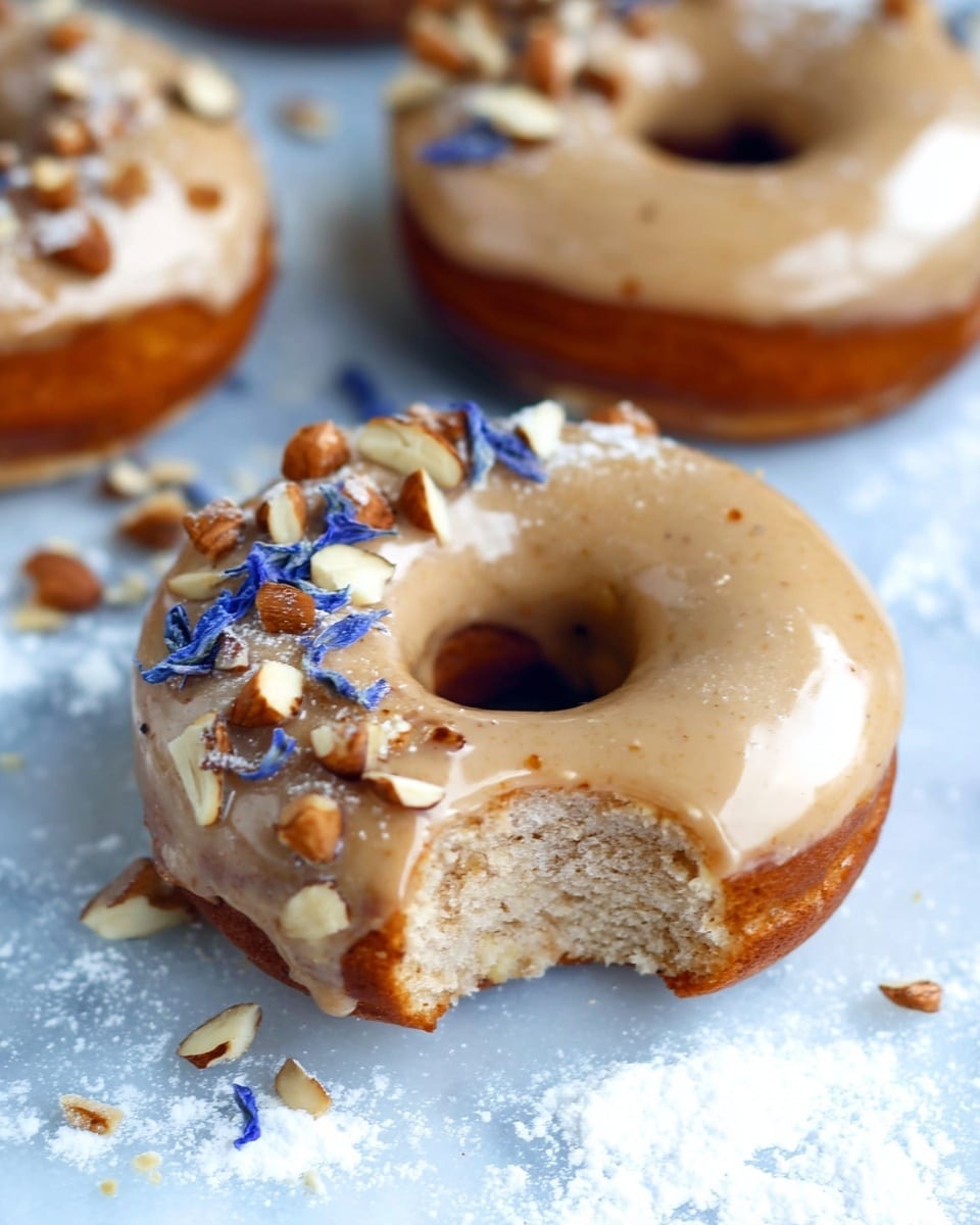 The image shows a close-up of a single donut with a smooth, light brown glaze covering it fully. On one side of the donut, there are small pieces of chopped nuts and delicate blue flower petals arranged in a neat pile. The donut has a soft, textured inside visible through a bite taken from one side, revealing a light brown crumb. The donut is placed on a white marbled surface with a light dusting of powdered sugar around it, and in the background, there are other glazed donuts slightly out of focus. photo taken with an iphone --ar 4:5 --v 7
