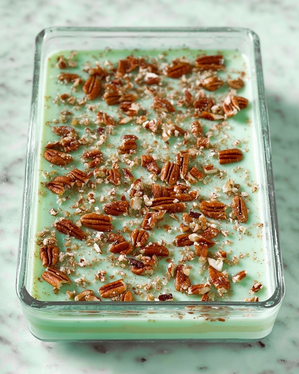 A clear rectangular glass dish holds a dessert with two visible layers: a smooth light green top layer with a creamy texture and a base layer beneath that is not visible. The top layer is sprinkled evenly with roughly chopped pecan nuts, giving a contrast of brown pieces on the green surface. The dish is placed on a white marbled surface, highlighting the colors of the dessert. Photo taken with an iphone --ar 4:5 --v 7