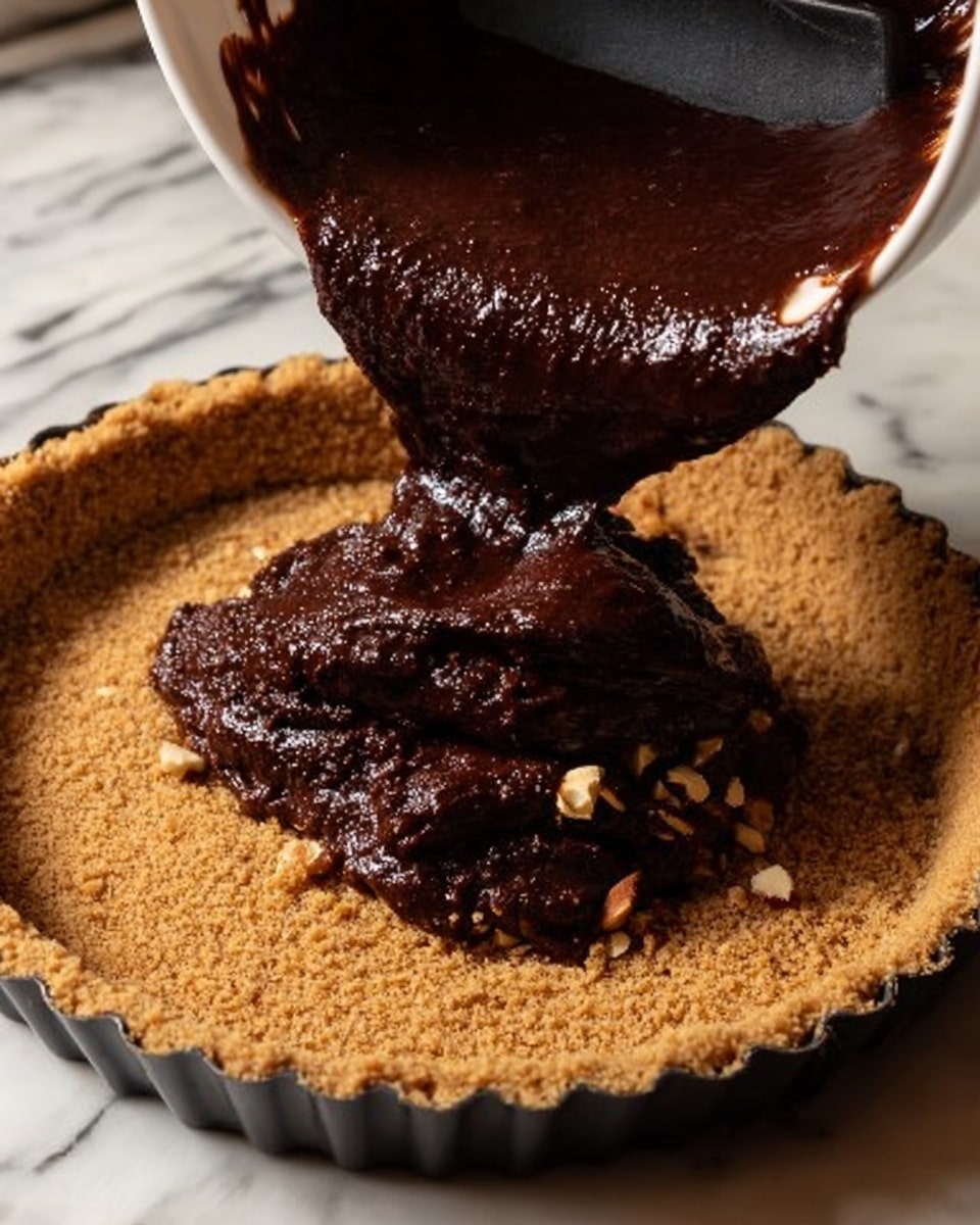 A close-up view of a cheesecakelike dessert in the making, showing a golden brown crumbly crust layer as the base inside a round tart pan. Thick, dark chocolate batter with a rich, glossy texture is being poured onto the crust from a white bowl held over it, with small nut pieces visible in the batter. A black silicone spatula is in the bowl helping to scoop out the batter. The background is a white marbled surface. Photo taken with an iphone --ar 4:5 --v 7