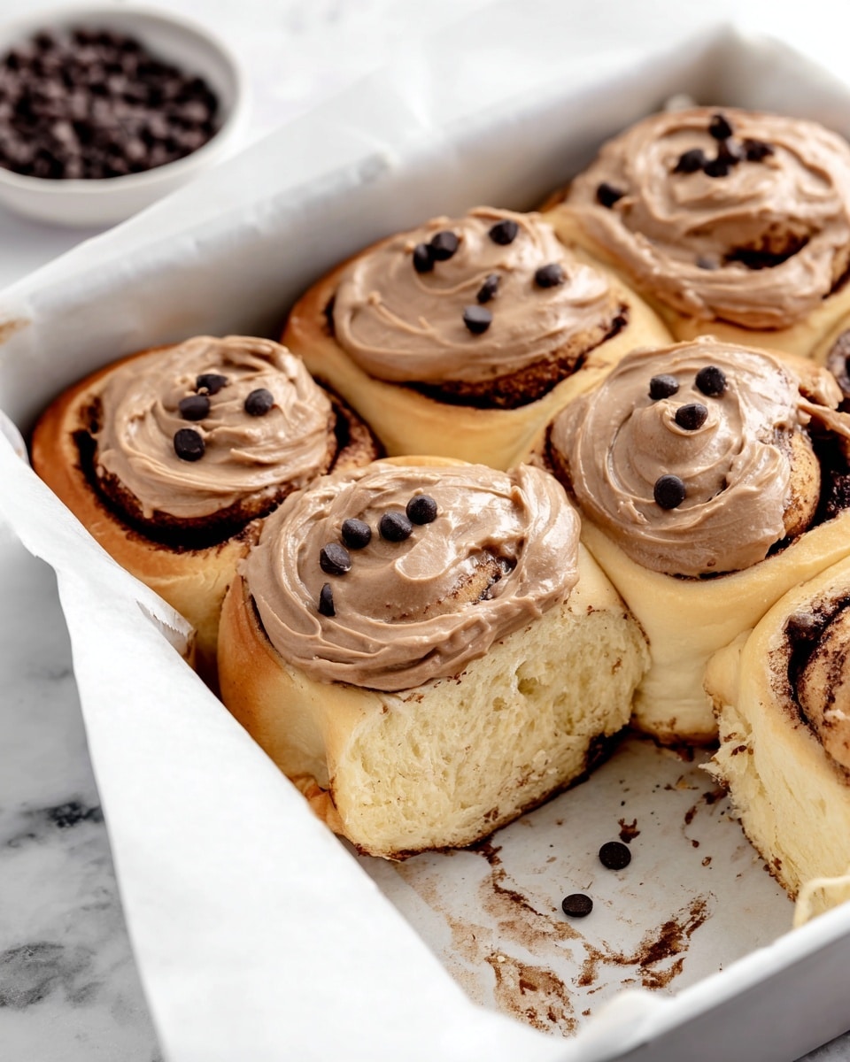 The image shows six soft cinnamon rolls inside a white baking pan lined with white parchment paper. Each roll has two layers: a light beige fluffy dough base with swirls of dark brown cinnamon filling, topped with a smooth light brown frosting spread thickly on top. There are small, round dark chocolate chips scattered on the frosting of each roll. One roll is missing from the bottom right corner, leaving visible cinnamon residue on the paper. In the background, there is a small white bowl filled with dark chocolate chips on a white marbled surface. Photo taken with an iphone --ar 4:5 --v 7
