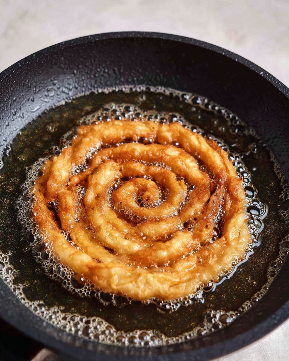 A round curly shape of golden brown batter is frying in dark oil inside a black textured frying pan. The batter is layered in spirals starting from the center and expanding outward with uneven edges, showing a crispy texture forming. Small bubbles from frying oil surround the swirl on the pan’s flat surface. The background is a white marbled texture with soft lighting. Photo taken with an iphone --ar 4:5 --v 7