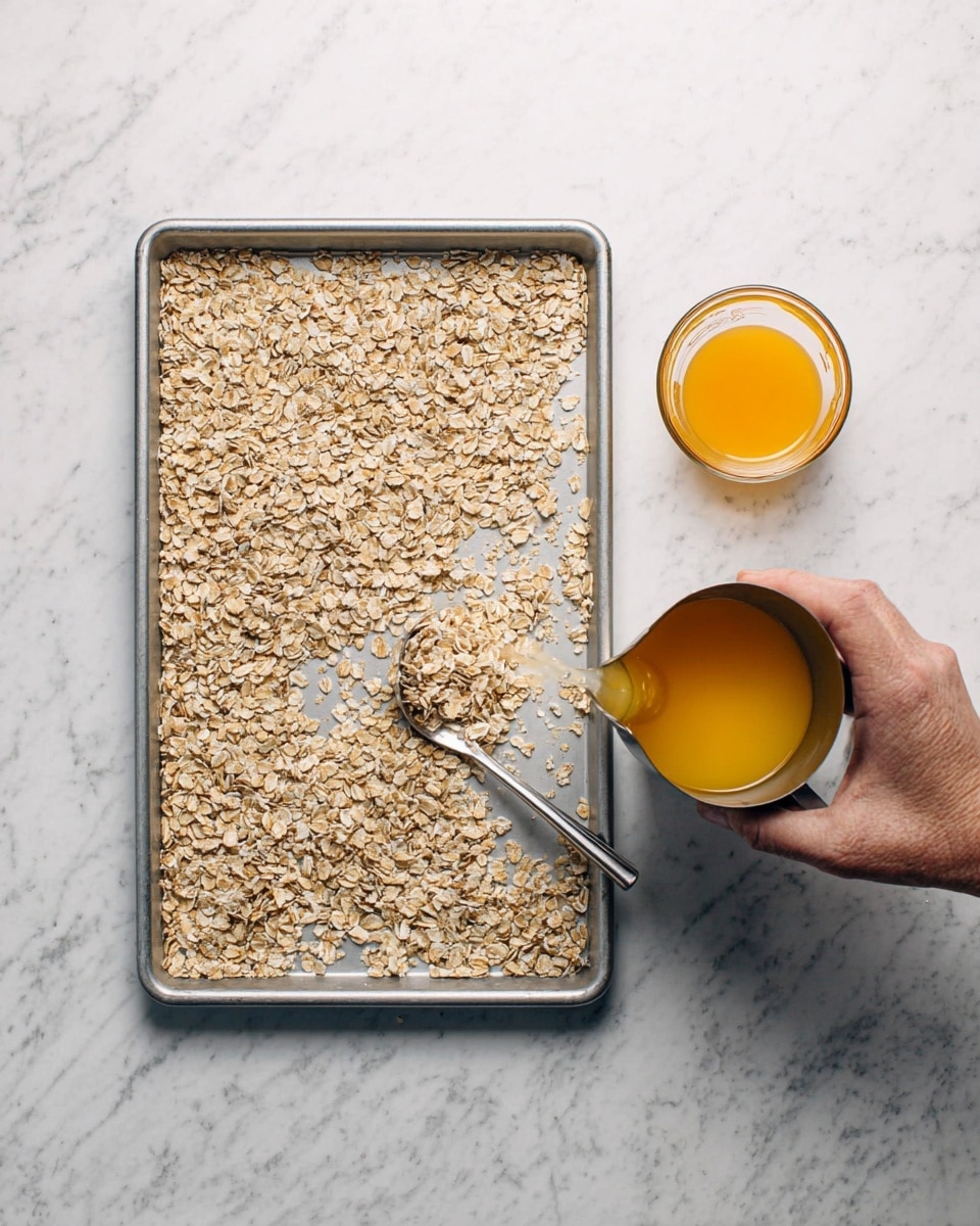 A top view of a rectangular metal baking tray lined with oats spread unevenly on a white marbled surface. On the right side of the tray, a woman's hand pours a liquid from a metal measuring cup, next to a glass measuring cup filled with orange liquid. The background is a smooth white marbled texture. Photo taken with an iphone --ar 4:5 --v 7