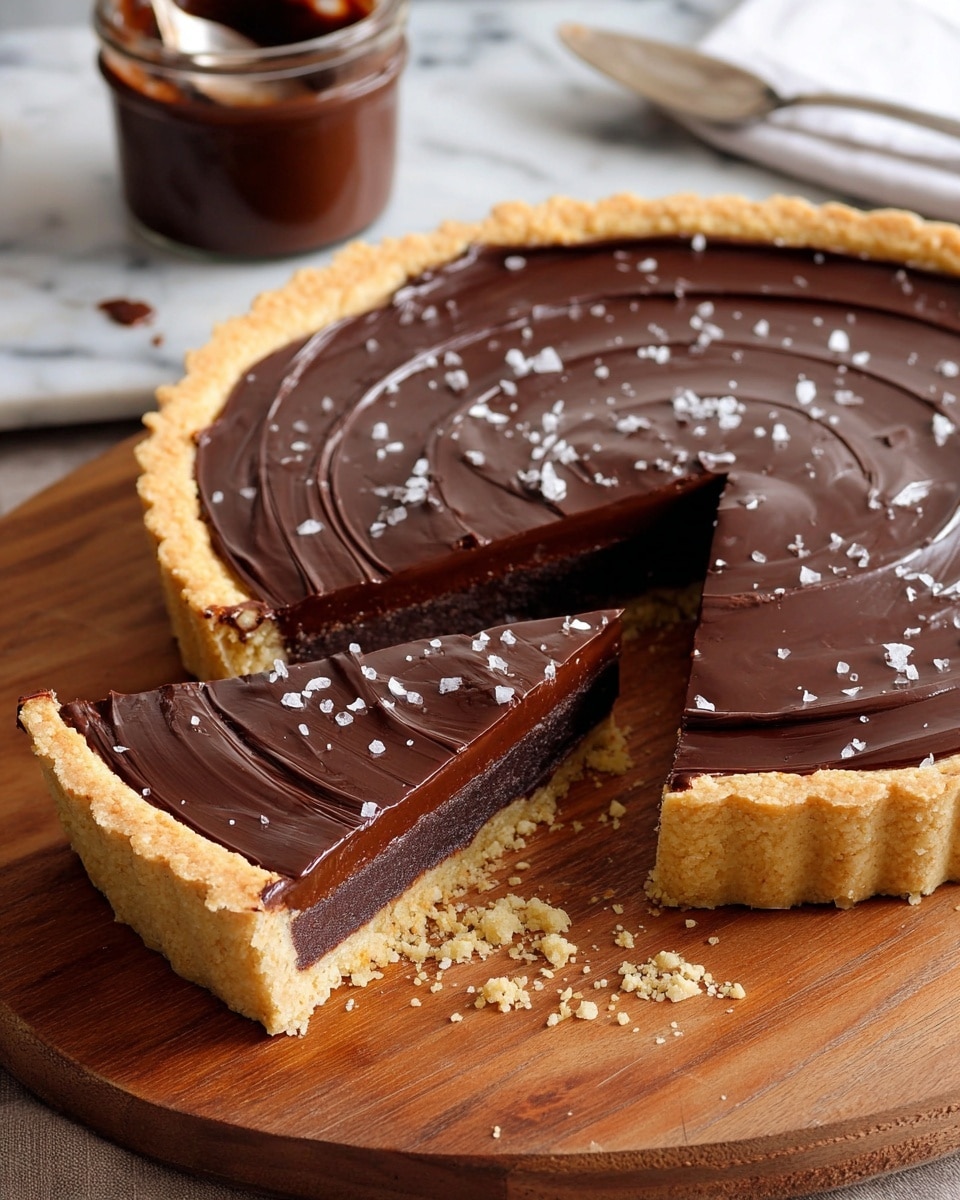 A round tart with one slice removed shows three layers: a light golden crumbly crust at the base and edges, a thick middle layer of dense dark chocolate filling, and a smooth glossy dark chocolate top layer with gentle swirl patterns. The top is sprinkled lightly with coarse salt flakes. The tart sits on a wooden board with some crumbs around it, and in the background, there is a white marbled surface with a clear glass jar of dark chocolate spread and a spoon. Photo taken with an iphone --ar 4:5 --v 7