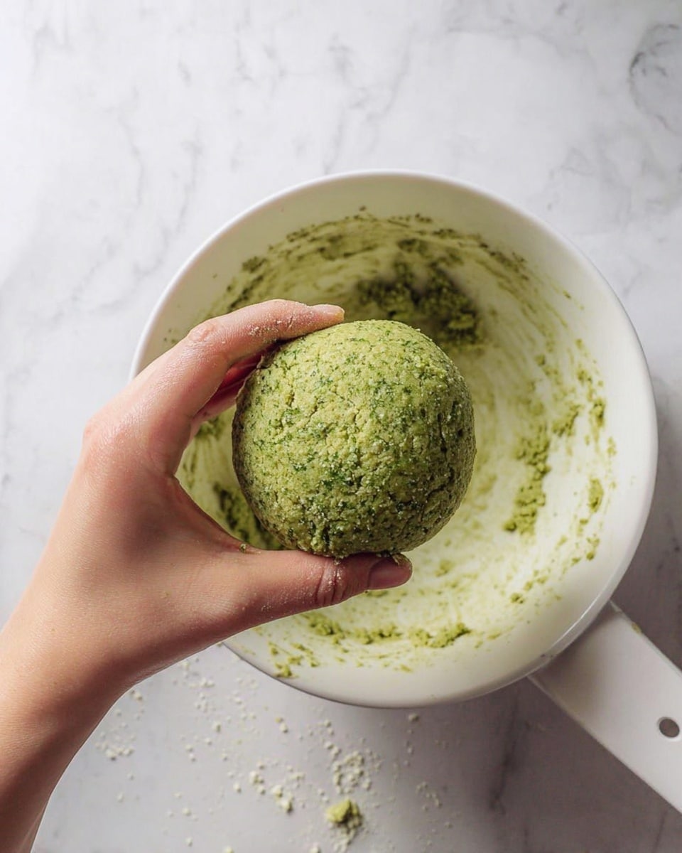 A woman's hand is holding a round ball of green dough with a rough texture, showing some small cracks on its surface. The dough looks moist and crumbly, with specks of lighter and darker green throughout. The hand is held above a white bowl that has green dough remnants smeared on the inside walls, indicating the dough was mixed there. The bowl sits on a white marbled surface with some dough bits scattered around it. A white spoon handle is visible on the right side of the bowl. photo taken with an iphone --ar 4:5 --v 7