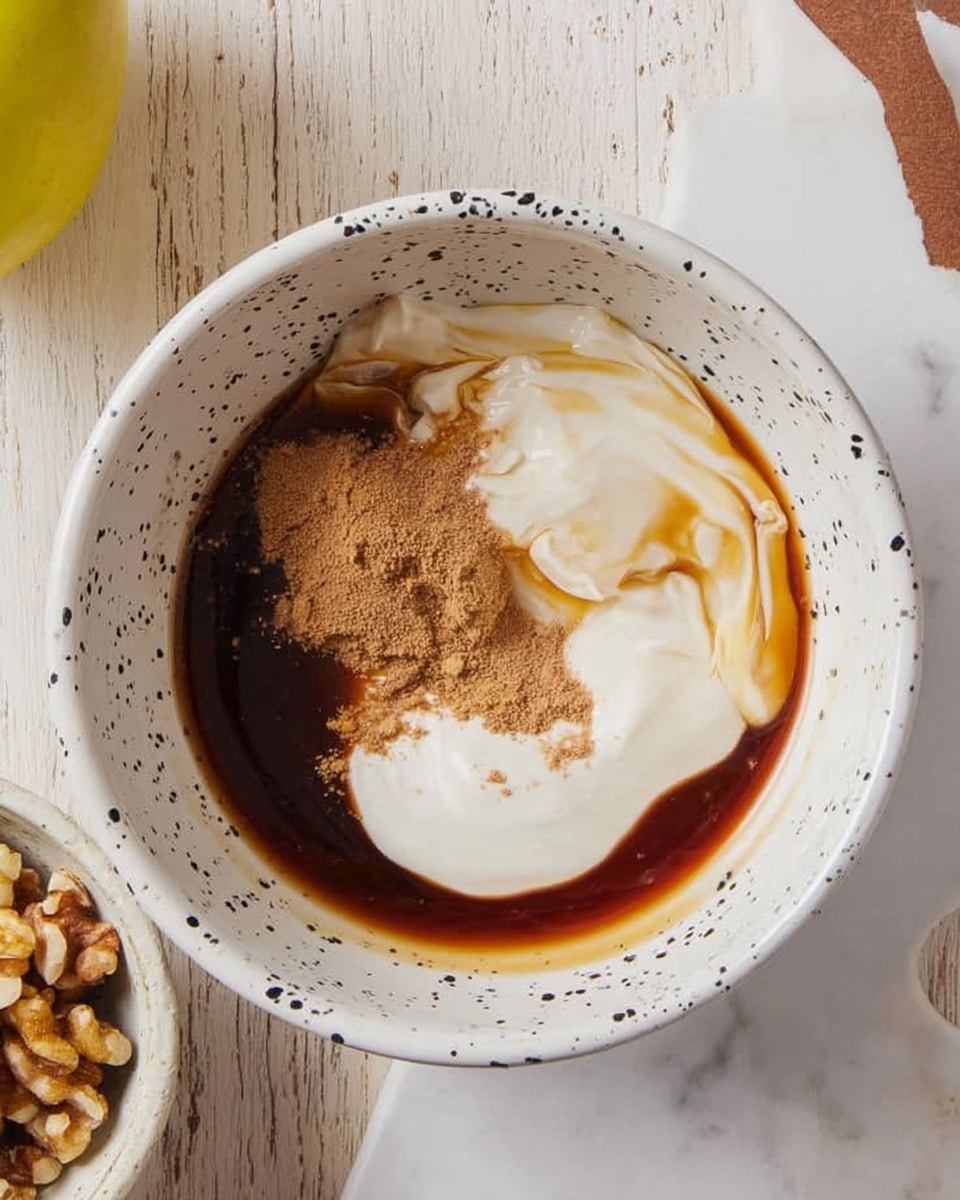 A white bowl with black speckles contains a three-layer mixture placed on a white marbled surface. The bottom layer is a dark brown syrup, pooling unevenly around the edges. On top, there is a smooth white creamy layer slightly swirled into the syrup. Next to the white cream, there is a small pile of light brown powder layered in a clumped shape. To the left, part of a yellow apple and a white speckled bowl with chopped walnuts are visible. Photo taken with an iphone --ar 4:5 --v 7