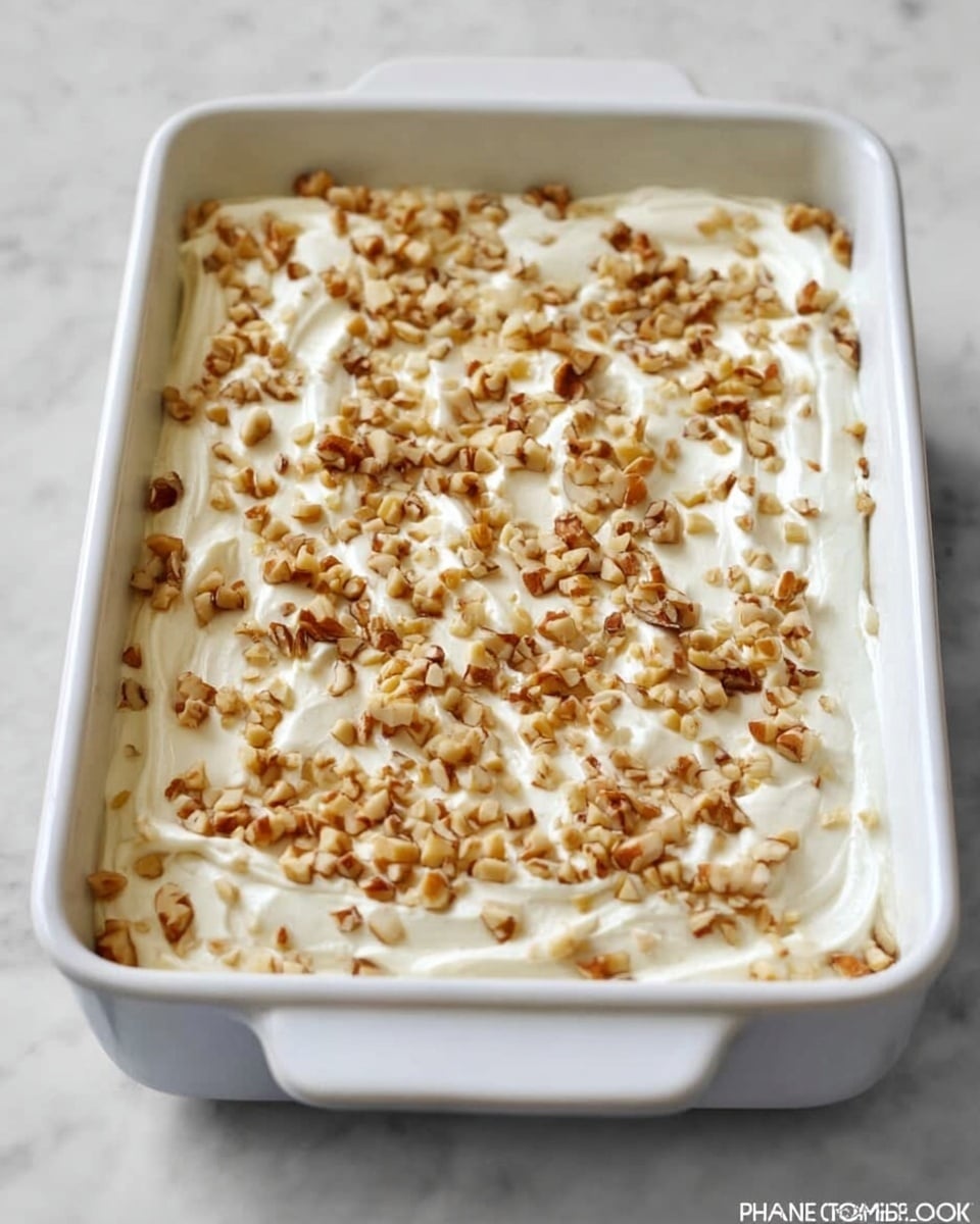 A white rectangular baking dish holds a creamy dessert spread evenly across the surface, topped with a scattered layer of small chopped nuts that add texture and light brown color contrast. The creamy layer is smooth with slight swirls and is a soft white color, covering the entire dish. The dish rests on a white marbled surface, enhancing the clean and simple presentation. Photo taken with an iphone --ar 4:5 --v 7