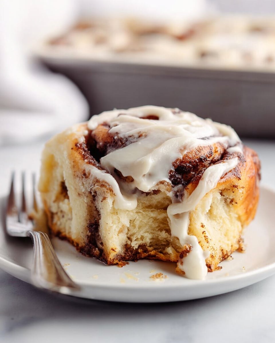 A soft cinnamon roll with visible thick swirls of brown cinnamon filling spread evenly in two layers inside its light golden dough, topped with a shiny white cream cheese frosting that drips slightly down the sides showing uneven texture. The roll is partly eaten and placed on a white plate against a white marbled surface background, with a silver fork on the left side and a blurred baking pan in the back. Photo taken with an iphone --ar 4:5 --v 7