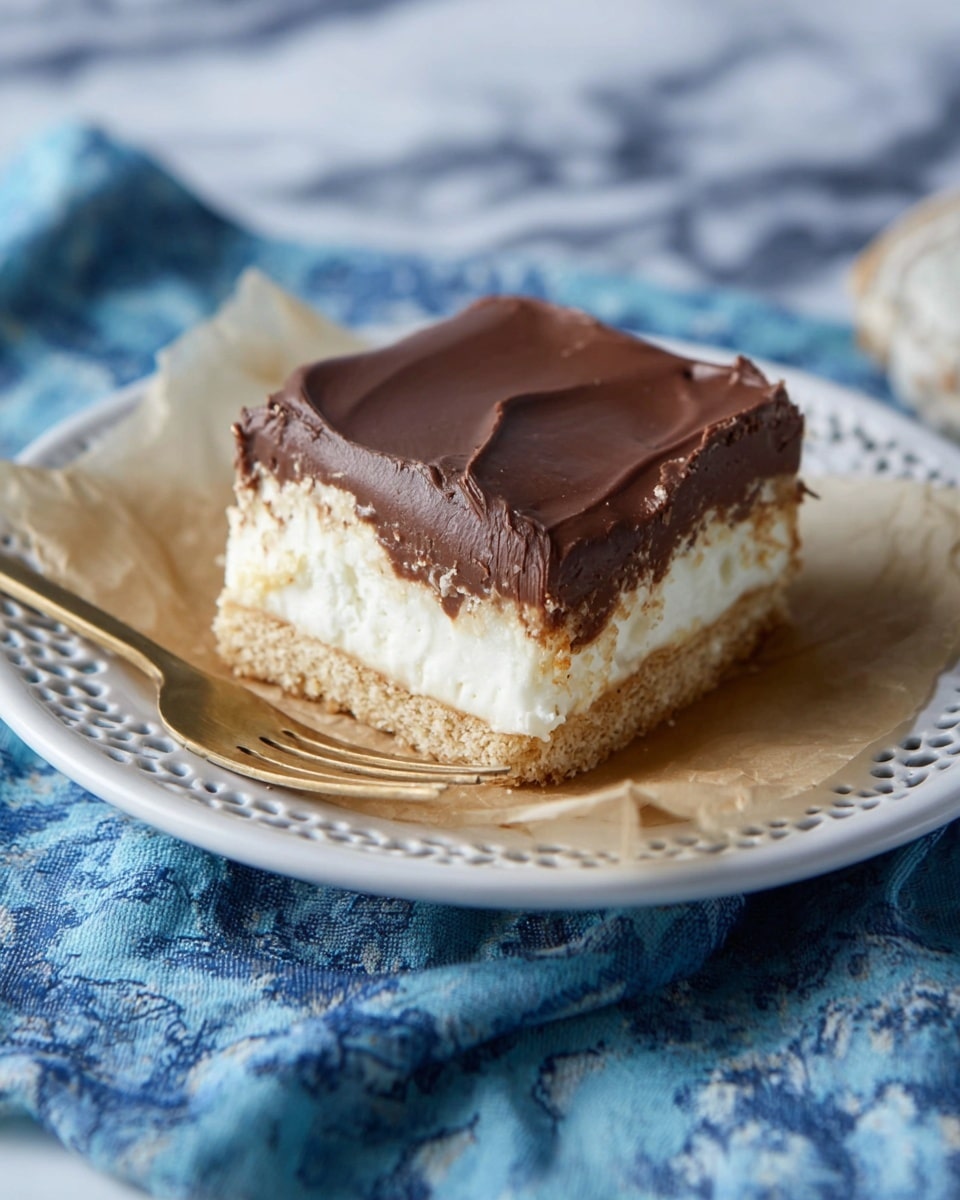 A square dessert bar with three visible layers sits on a piece of parchment paper on a white, lace-edged plate. The bottom layer is light brown and crumbly like a biscuit base, the middle layer is thick and creamy white, and the top layer is smooth, thick dark brown chocolate spread evenly. A small bronze fork lies next to the dessert on the plate. The plate rests on a blue fabric with white patterns, and the background is a white marbled texture. Photo taken with an iphone --ar 4:5 --v 7