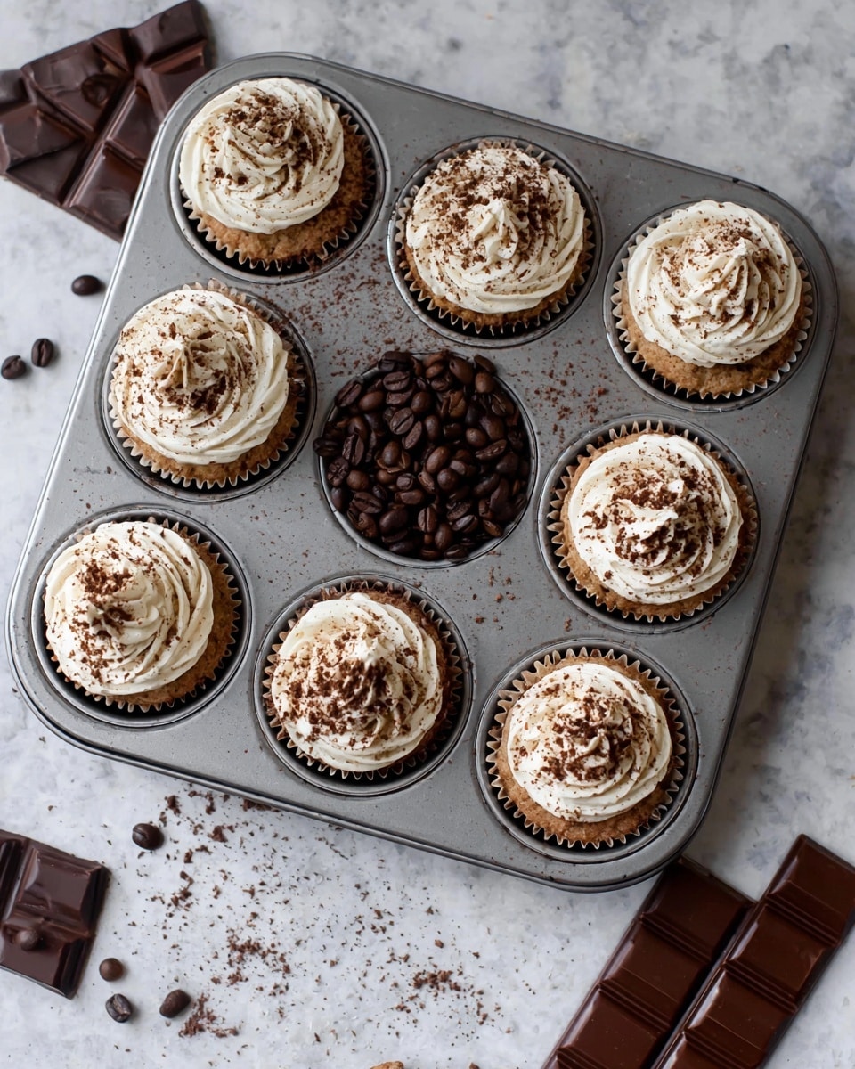 A metal cupcake pan holds eight cupcakes and one cup filled with dark brown coffee beans in the center. Each cupcake has one layer of light brown cake, topped with a swirl of white cream frosting sprinkled with fine dark brown cocoa powder. The pan sits on a white marbled surface scattered with a few coffee beans and small chocolate pieces. Pieces of dark chocolate bars rest nearby to the right of the pan. Photo taken with an iphone --ar 4:5 --v 7