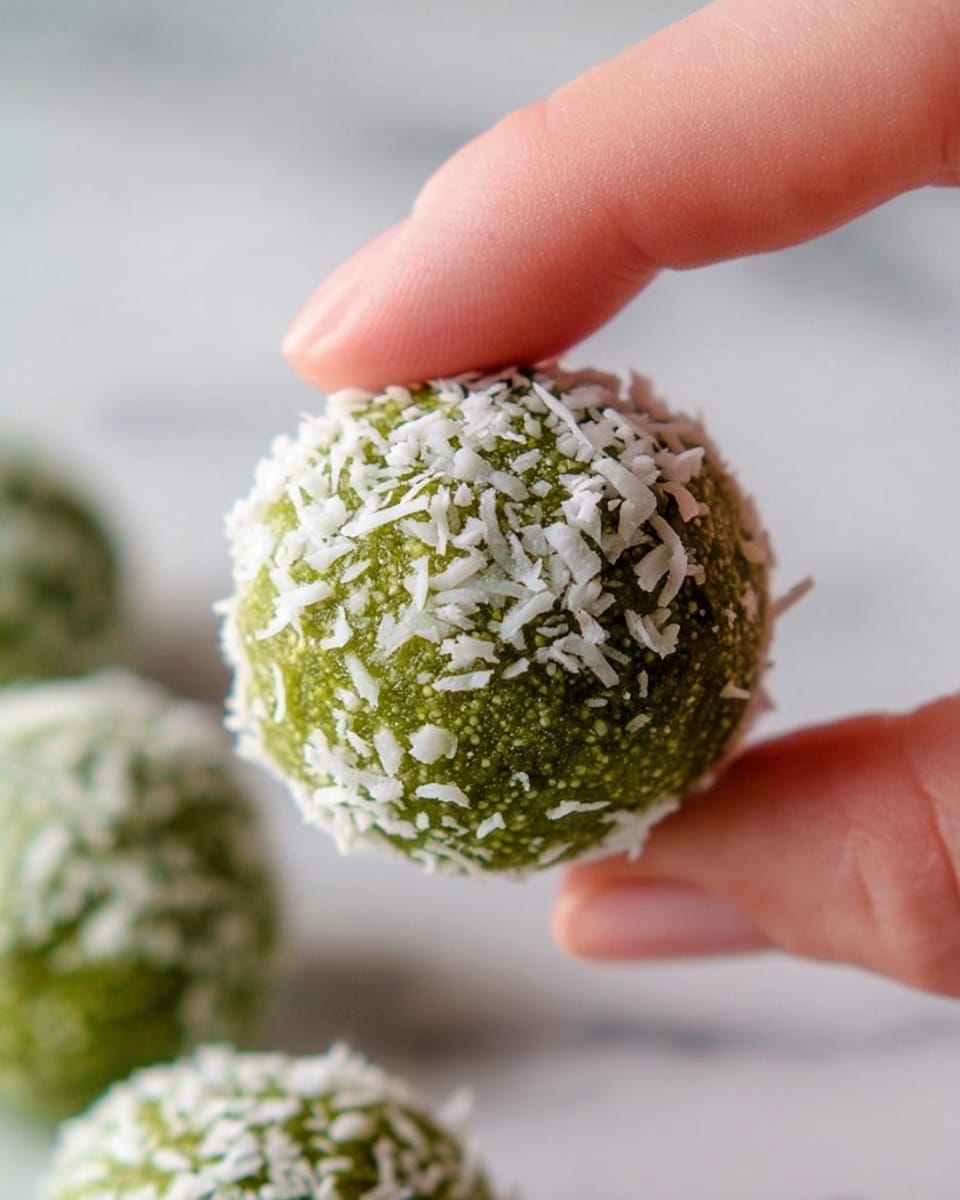 A close-up shows a round green ball covered with small white coconut flakes. The ball has a smooth, slightly shiny green base with textured coconut shreds sticking out all over. A woman's hand is holding the ball between the thumb and index finger. Below, several similar green balls covered in coconut flakes sit on a white marbled surface, slightly out of focus. The overall light is soft and natural, highlighting the white flakes and the green color of the ball photo taken with an iphone --ar 4:5 --v 7