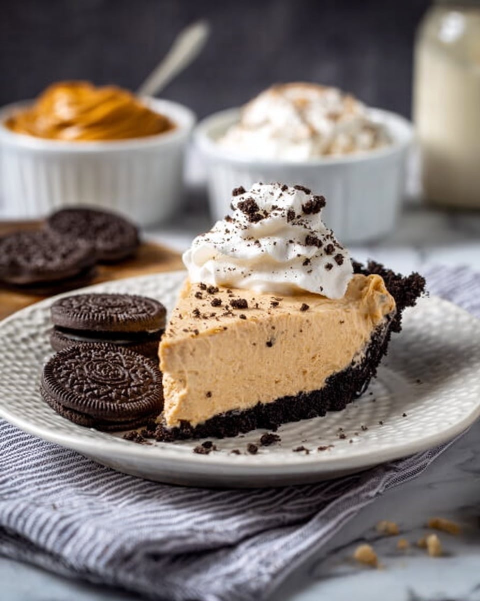 A slice of light brown creamy pie with a thick dark brown crumb crust on the bottom sits on a white textured plate, topped with a dollop of white whipped cream sprinkled with small dark cookie crumbs; two dark sandwich cookies lean on the left side of the pie slice. In the background, two white bowls hold creamy peanut butter and whipped cream, placed on a white marbled surface with a striped gray and white cloth to the right. Photo taken with an iphone --ar 4:5 --v 7