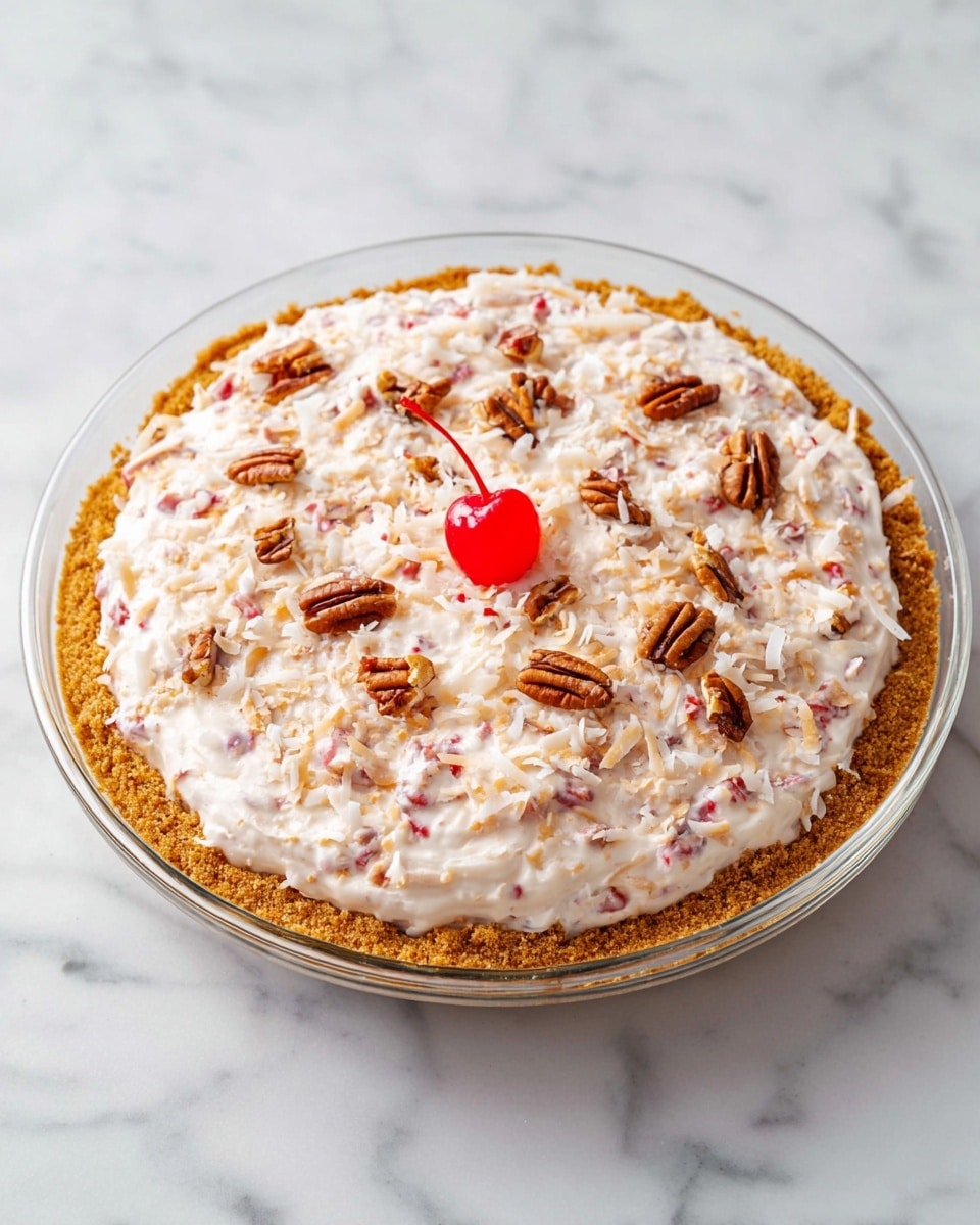 A round dessert sits in a clear glass pie dish on a white marbled surface, showing three main layers. The bottom layer is a golden-brown crust with a crumbly texture. On top is a thick, creamy white layer mixed with small red pieces and sprinkled with shredded coconut, giving it a fluffy, textured look. The top is decorated with scattered small brown pecan nuts and a single bright red cherry placed in the center. Photo taken with an iphone --ar 4:5 --v 7