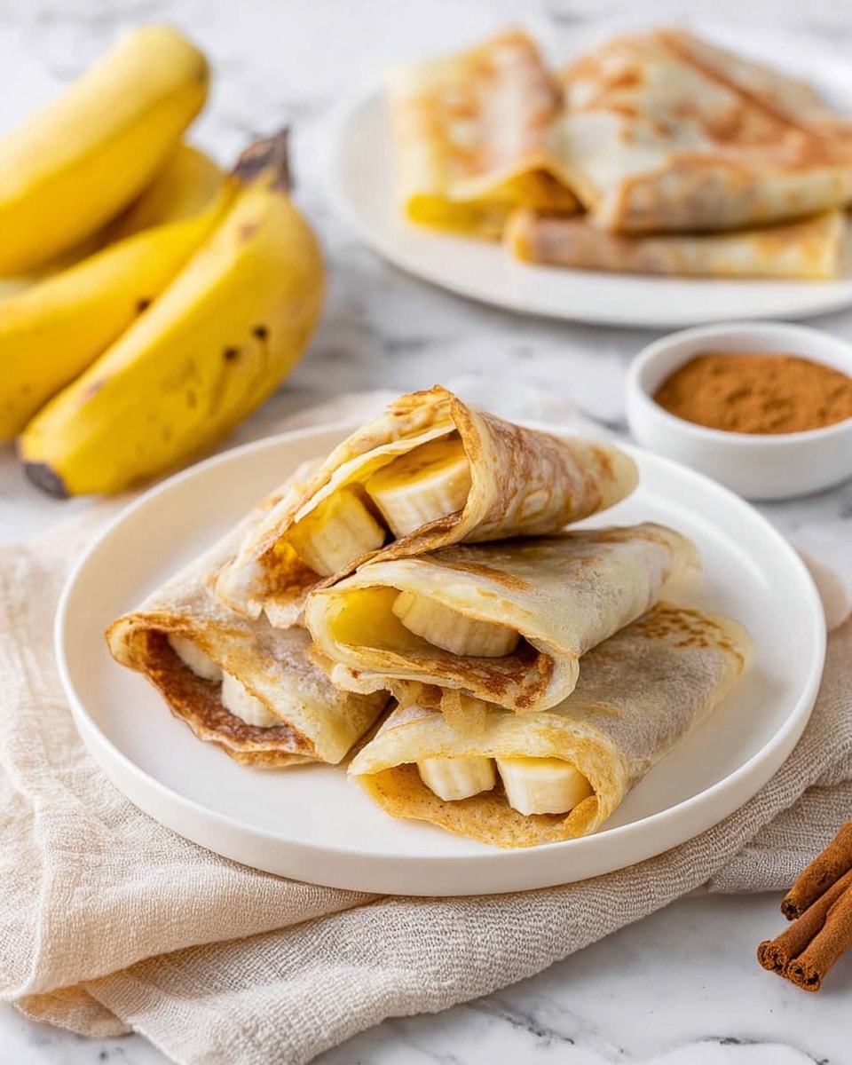 The image shows a white plate holding several golden-brown folded crepes with visible yellow banana slices and light brown filling inside. On top, three pieces are open to show the soft banana layer inside the thin, slightly toasted crepe. In the background, another white plate with more folded crepes can be seen, along with two whole yellow bananas to the left and a small white bowl with brown cinnamon powder at the bottom right. The scene sits on a white marbled surface with a light beige cloth under the front plate. Photo taken with an iphone --ar 4:5 --v 7