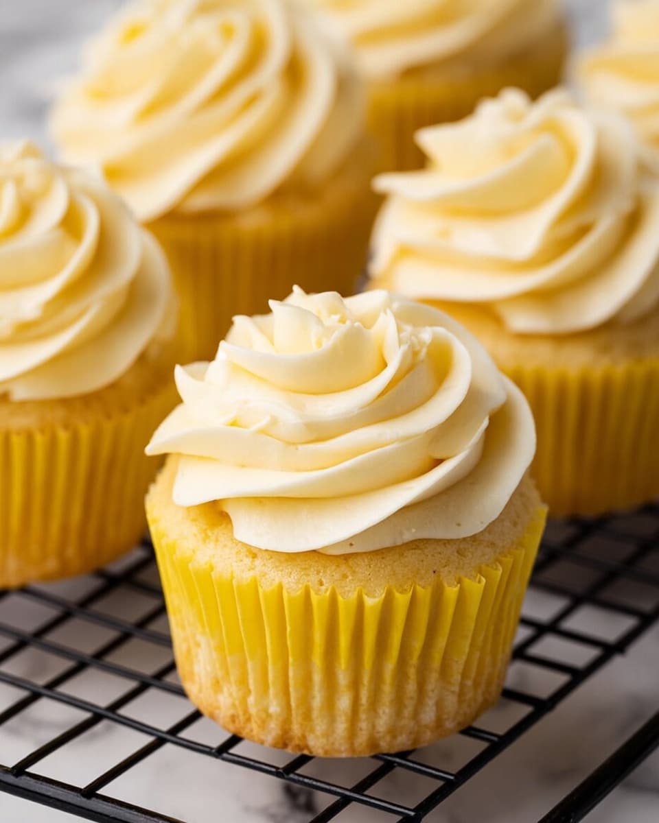 The image shows several yellow cupcakes with one in the center in focus. Each cupcake has two main layers: a yellow cake base wrapped in a yellow paper liner, topped with a thick swirl of creamy light yellow frosting that has soft peaks and smooth texture. The cupcakes are placed on a black metal cooling rack with a close-up view that highlights the frosting's swirled details and the cake's soft crumb texture. The background is a white marbled surface. photo taken with an iphone --ar 4:5 --v 7
