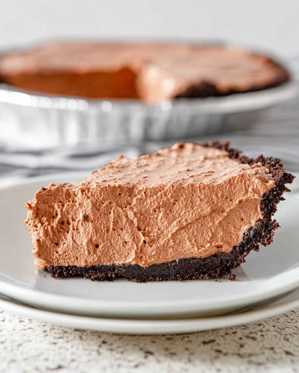 The image shows a thick slice of chocolate mousse pie on a white plate with a dark chocolate crust at the bottom. The mousse layer is light brown and fluffy, filling most of the slice with a creamy yet airy texture. The crust appears crumbly and dark, contrasting the soft mousse on top. In the background, the pie from which the slice was taken is visible, with a shiny, smooth mousse surface and the crust visible on the sides. The setting has a white marbled texture under the plate and a soft light highlighting the pie's texture. photo taken with an iphone --ar 4:5 --v 7