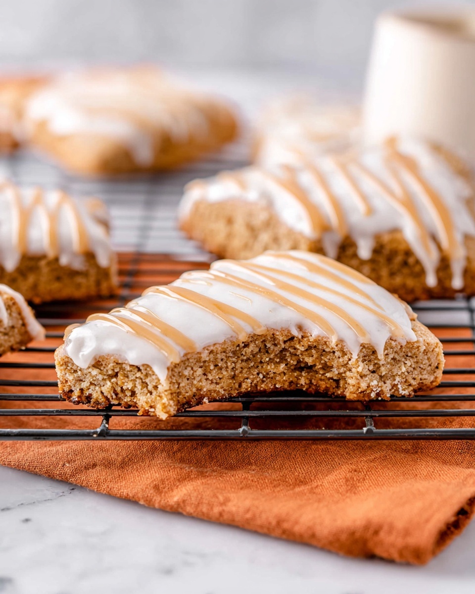 Gluten-Free Pumpkin Scones Recipe 4 The image shows two triangular pastries on a metal cooling rack over an orange cloth, all set on a white marbled surface. Each pastry has a brown, slightly textured base with a smooth layer of white icing covering the top. Over the white icing, there are zigzag stripes of light brown glaze. One of the pastries has a bite taken out of it, revealing the dense, crumbly inside. In the background, more of these pastries are softly blurred. photo taken with an iphone --ar 4:5 --v 7