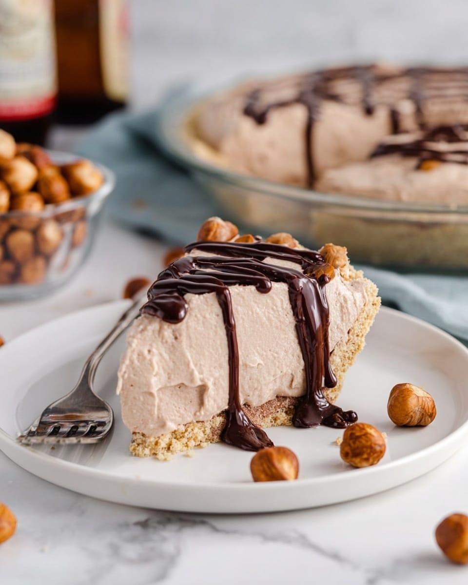 The image shows a slice of light brown mousse pie on a white plate with a crumbly beige crust layer at the bottom. The mousse layer is thick and smooth, and the slice is drizzled with dark chocolate sauce on top, running down the sides. Around the slice, there are several whole hazelnuts scattered on the white marbled surface. A silver fork rests on the plate next to the pie slice. In the background, there is a larger mousse pie in a clear round dish with more chocolate drizzle on top and a glass bowl filled with hazelnuts. A blurred bottle is also visible, suggesting a drink. photo taken with an iphone --ar 4:5 --v 7