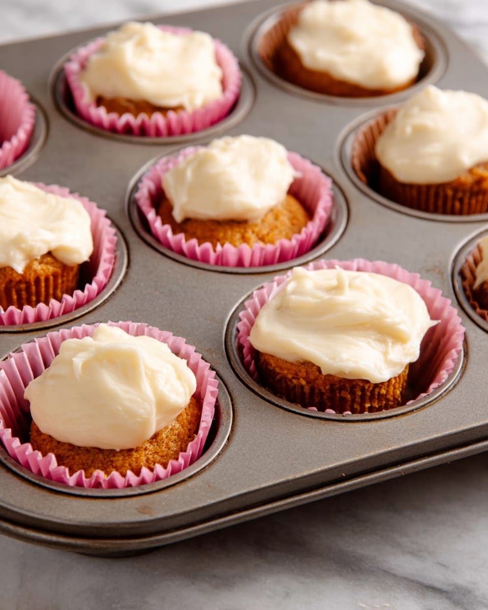 A close-up view of a metal muffin tray containing six baked cupcakes, each lined with a pink paper cup. The cupcakes have two layers visible: a bottom layer that is a dense, moist orange-brown cake, and a top layer of creamy, smooth white frosting dolloped unevenly on each cupcake. The tray is placed on a white marbled surface, adding a clean, bright contrast to the warm colors of the cupcakes. photo taken with an iphone --ar 4:5 --v 7
