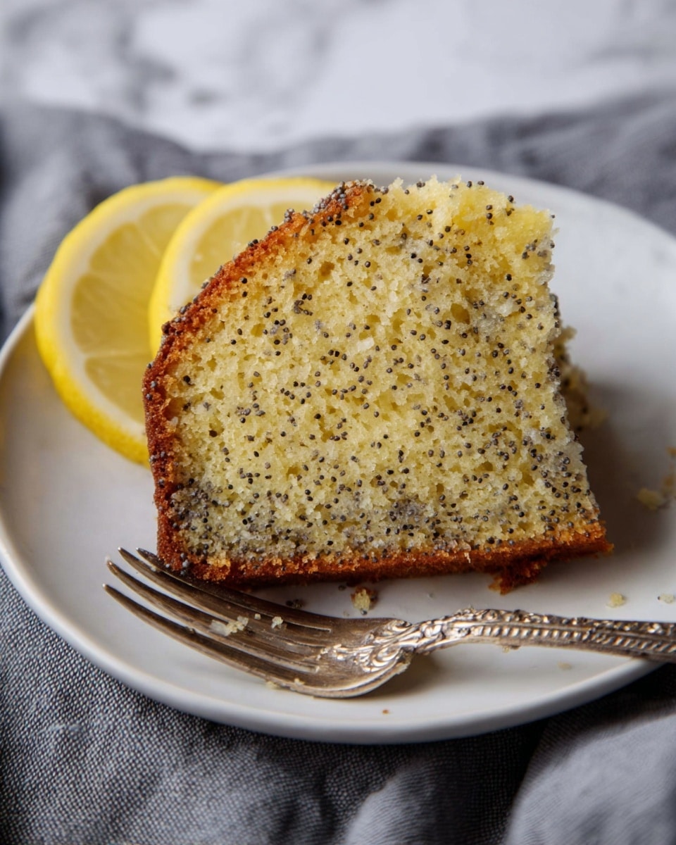 A single slice of lemon poppy seed cake with a light yellow crumb speckled with small black poppy seeds and a golden brown crust sits on a white plate. Two thin lemon wedges with bright yellow rinds rest behind the cake slice. An ornate silver fork is placed at the bottom of the plate. The plate is set on a soft grey cloth, and the background has a white marbled texture. Photo taken with an iphone --ar 4:5 --v 7