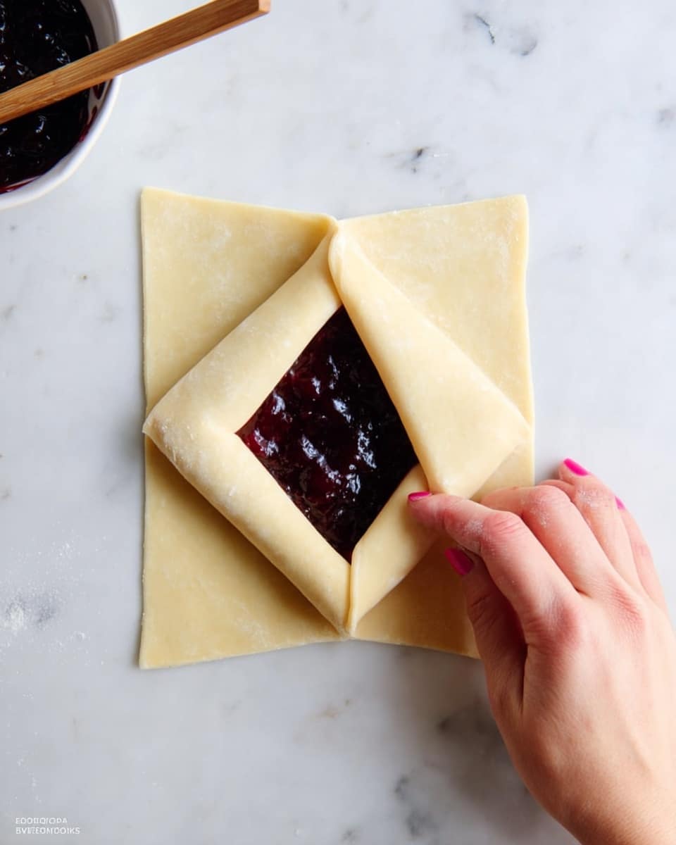 A woman's hand with pink nail polish is folding the sides of a pale yellow dough square over a dark purple, thick jam filling in the center. The dough is light, smooth, and thin, shaped like an envelope with layers folded over the jam in the middle. The scene is set on a white marbled surface. At the top left edge, there is a white bowl with dark jam and a wooden chopstick inside. Photo taken with an iphone --ar 4:5 --v 7