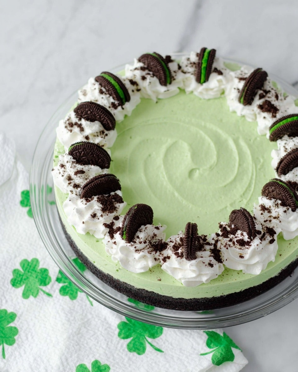 The image shows a round dessert with three main layers. The bottom layer is a crushed dark cookie crust. Above it is a thick, smooth, light green layer of creamy filling that covers the entire surface evenly with a slight swirl texture. The top edge is decorated with a ring of white whipped cream dollops, each topped with a halved dark chocolate cookie with a green filling visible. Crushed dark cookie crumbs are sprinkled over the green layer near the whipped cream, adding texture. The dessert sits on a clear glass plate placed on a white marbled surface with a white cloth featuring green shamrocks under one side of the plate. Photo taken with an iphone --ar 4:5 --v 7