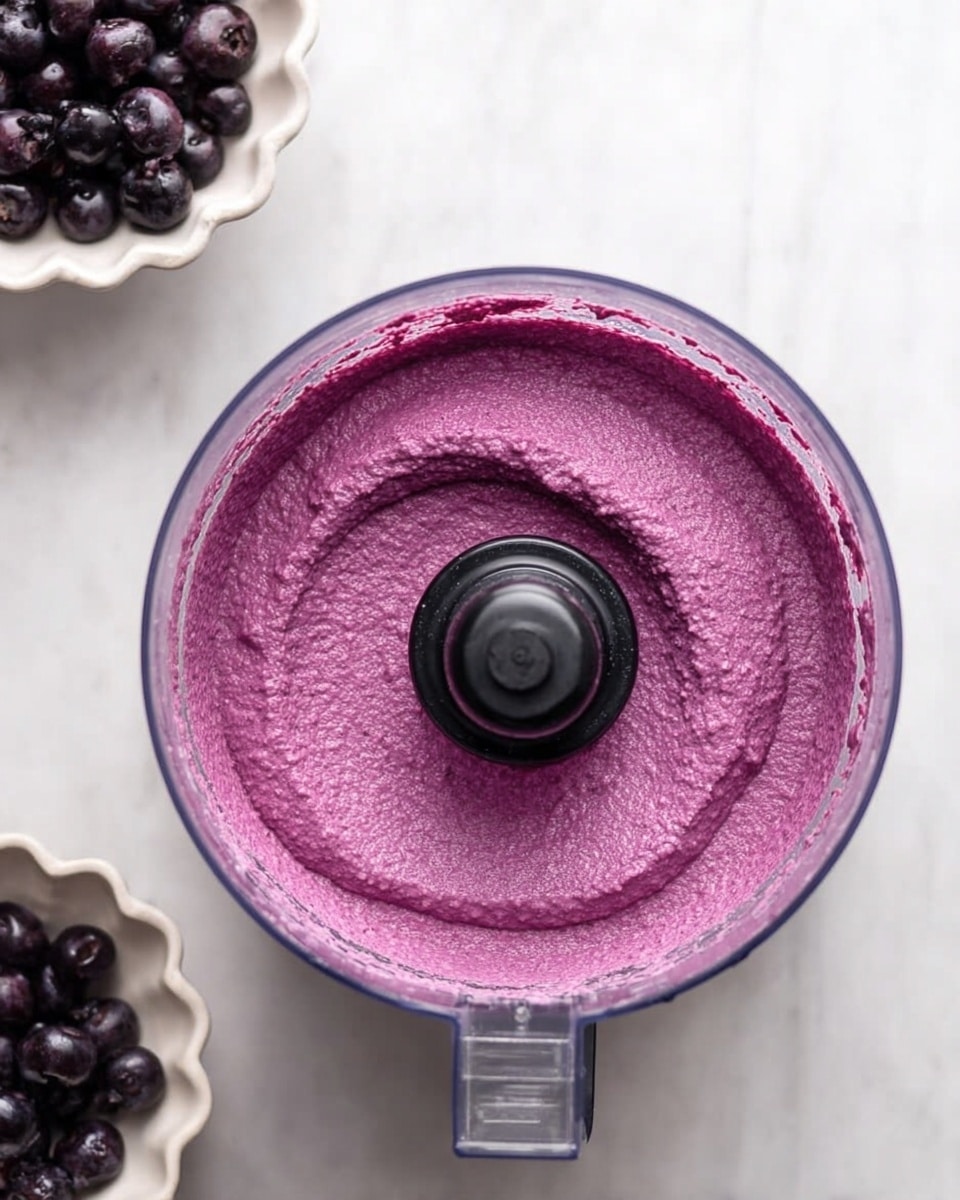 The image shows a close-up top view of a clear food processor bowl filled with a smooth, thick purple mixture that has a slightly textured surface. The mixture is evenly spread and swirled slightly around the central black blade base in the middle of the bowl. To the top left of the bowl, there is a small white scalloped bowl filled with whole dark purple berries. The whole scene is set on a white marbled surface. photo taken with an iphone --ar 4:5 --v 7