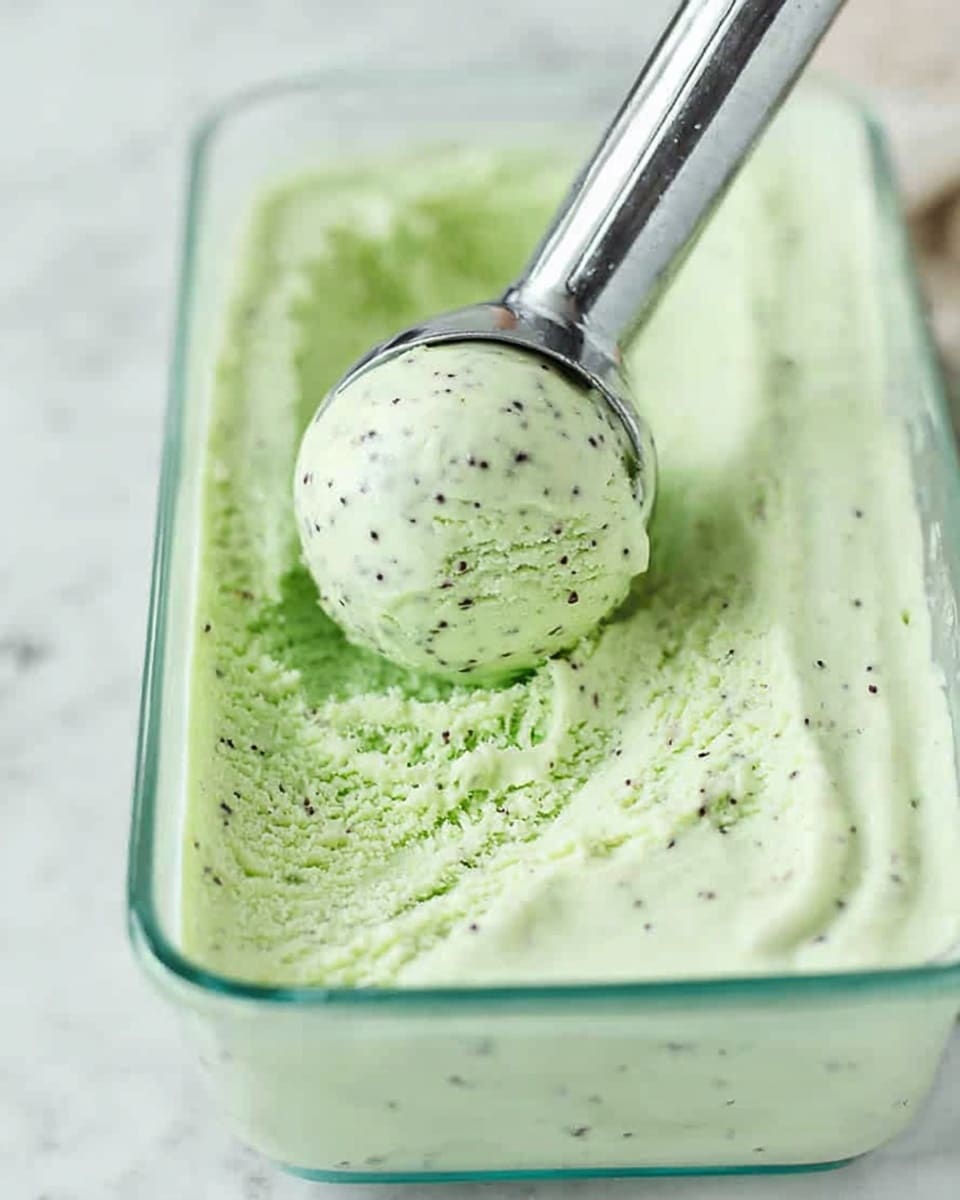 A close-up of a clear glass rectangular container filled with pale green ice cream that has small dark specks evenly spread throughout. In the center, a shiny silver ice cream scoop is lifting a smooth, round ball of the same pale green ice cream, showing its creamy texture. The container sits on a white marbled surface, creating a clean and bright background that contrasts softly with the ice cream's color. Photo taken with an iphone --ar 4:5 --v 7
