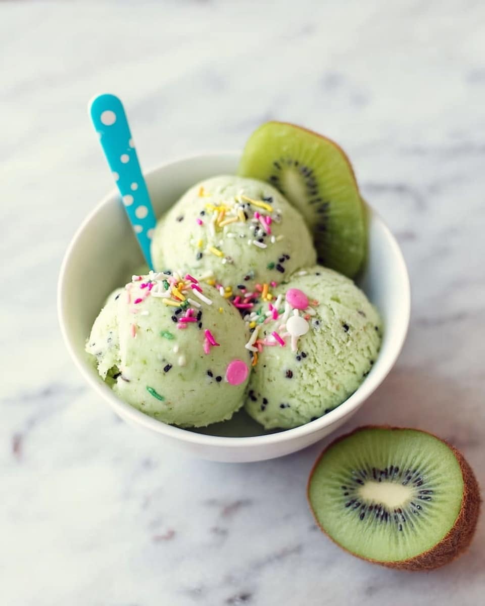A white bowl sits on a white marbled surface, filled with three round scoops of pale green kiwi-flavored ice cream with small black kiwi seeds visible throughout. On top of the ice cream scoops are small, colorful sprinkles in pink, yellow, white, and black. A thin slice of bright green kiwi fruit with a dark center is placed on the ice cream. A blue spoon with white polka dots rests inside the bowl. Next to the bowl is a halved kiwi fruit showing its vibrant green flesh and black seeds. Photo taken with an iphone --ar 4:5 --v 7