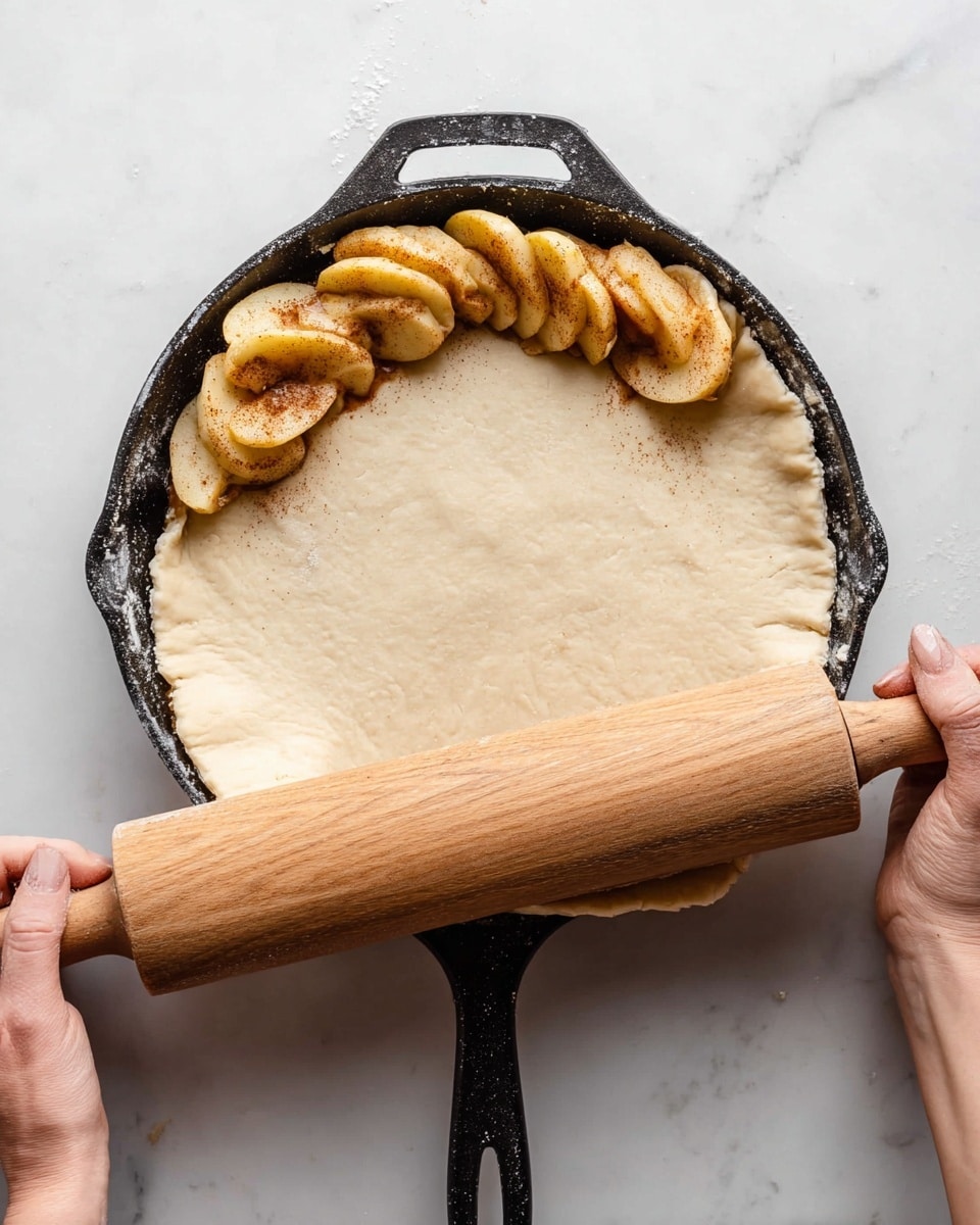 A black cast iron pan holds three halved apples sprinkled with cinnamon, arranged at the top inside edge. A light beige pie dough is being rolled out over the apples with a wooden rolling pin held by two woman's hands on either side, covering part of the apples. The dough has a soft, slightly flaky texture and extends over the pan edges in an uneven circle. The scene is set on a white marbled surface. photo taken with an iphone --ar 4:5 --v 7