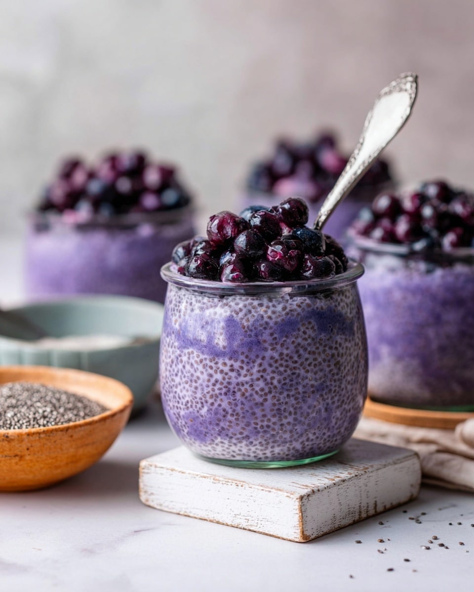 The image shows clear glass jars filled with three layers: the bottom layer is thick, purple chia pudding with visible small chia seeds throughout, the middle layer looks uniform and creamy purple, and the top layer is a mix of dark purple and black fresh blueberries piled generously. One jar has a silver spoon inside, breaking the smooth surface of the pudding. The jars are placed on and around a small white rustic wooden block, all set on a white marbled surface. In the background, there are bowls of chia seeds and more blueberries, slightly blurred. Photo taken with an iphone --ar 4:5 --v 7