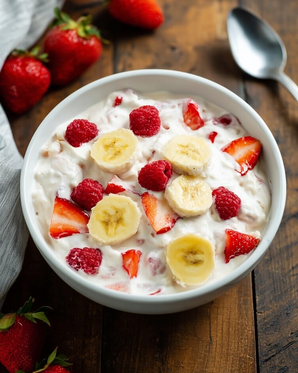 The image shows a white bowl filled with a creamy white mixture that looks soft and fluffy, with slices of yellow banana, bright red strawberries, and small red raspberries mixed in and resting on top. The fruit pieces are spread evenly throughout and on the surface, adding pops of red and yellow against the white cream. The bowl is placed on a wooden table, with some whole strawberries nearby and a silver spoon to the right. photo taken with an iphone --ar 4:5 --v 7