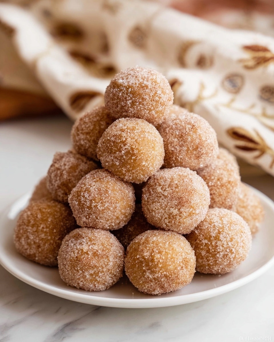 A white plate filled with about fifteen round balls stacked in a pyramid shape. Each ball has a rough texture and is covered in a light dusting of sugar and cinnamon, giving a sandy brown and slightly white appearance. The balls are uniform in size and closely packed together, showing granulated sugar crystals on their surface. The plate sits on a white marbled table, and a soft focus beige cloth with brown patterns is visible in the background. photo taken with an iphone --ar 4:5 --v 7