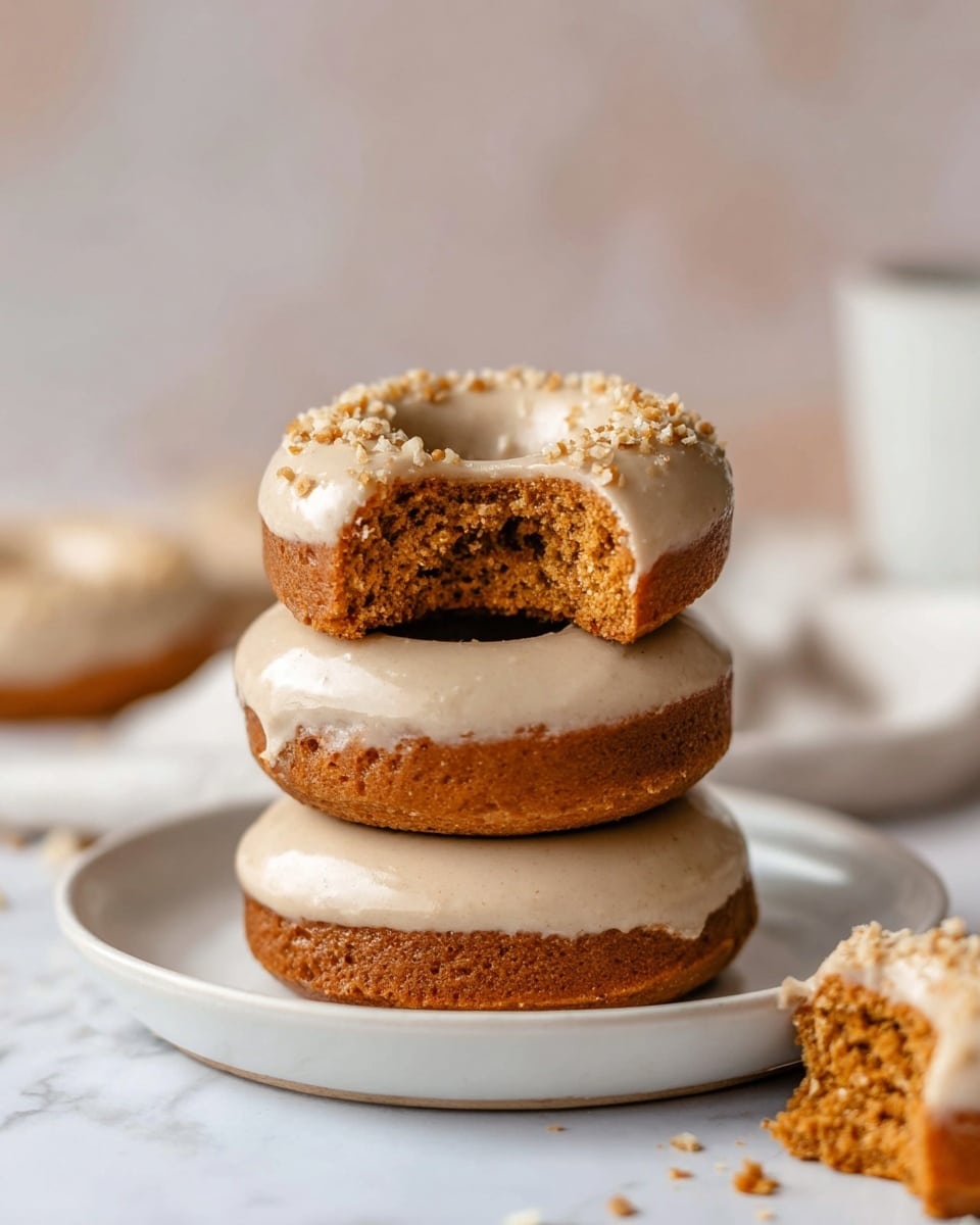 Three donuts are stacked on a white plate on a white marbled surface. Each donut has a thick layer of light beige frosting on top with small nut crumbs sprinkled over it. The top donut has a bite taken from it, revealing a soft, brown, cake-like inside. There is a small piece of a donut with the same frosting lying next to the plate. The background is softly blurred with light neutral tones. Photo taken with an iphone --ar 4:5 --v 7