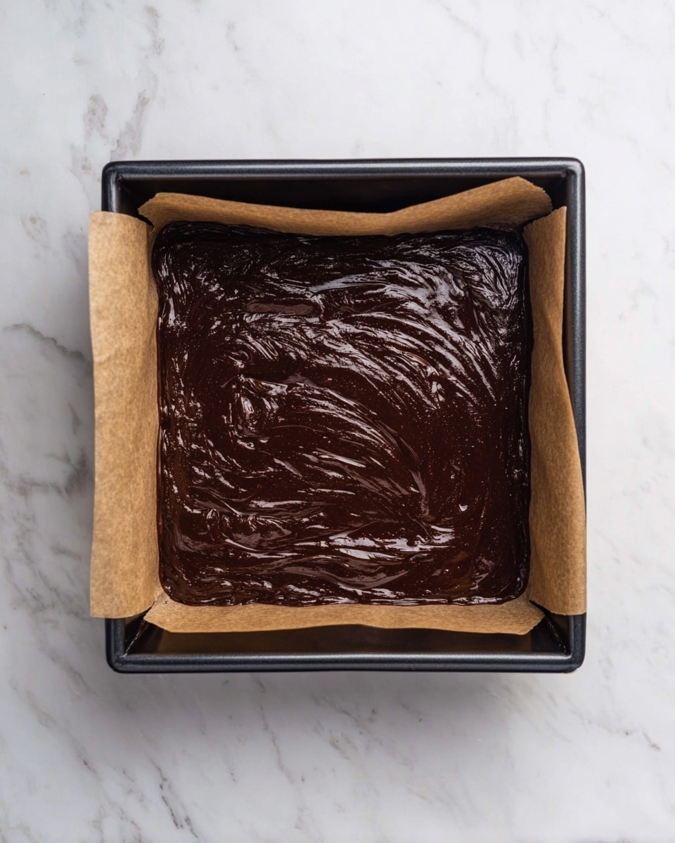The image shows a square black baking pan lined with brown parchment paper filled with dark brown, glossy chocolate batter spread evenly across the pan. The surface of the batter has a smooth, shiny texture with visible swirl patterns from mixing. The pan is placed on a white marbled surface. photo taken with an iphone --ar 4:5 --v 7