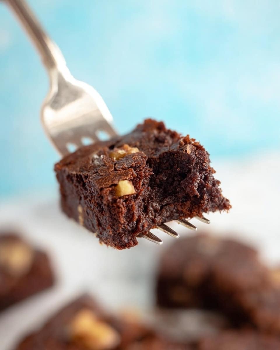 A small square piece of chocolate brownie is held on a silver fork close to the camera, showing its moist and dense texture. The top layer of the brownie is slightly crunchy with visible small pieces of nuts, while the inside looks soft and rich, dark brown in color. The background shows some blurred pieces of brownie resting on a white marbled surface with a soft focus blue area behind it. The image captures the details of the chocolate and nut textures clearly, emphasizing the delicious look of the brownie photo taken with an iphone --ar 4:5 --v 7