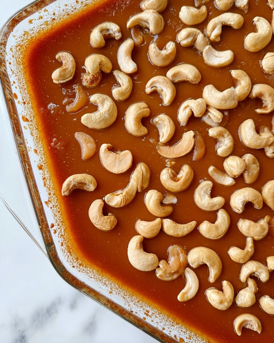 This image shows a close-up of a clear glass pan filled with a single layer of glossy caramel sauce, which is deep golden brown in color and smooth in texture. On top of the caramel, there is an even layer of whole cashew nuts scattered across the surface, their pale beige color contrasting with the rich caramel beneath. The edges of the pan reveal some caramel residue, adding a rustic look. The pan is placed on a white marbled surface. Photo taken with an iphone --ar 4:5 --v 7
