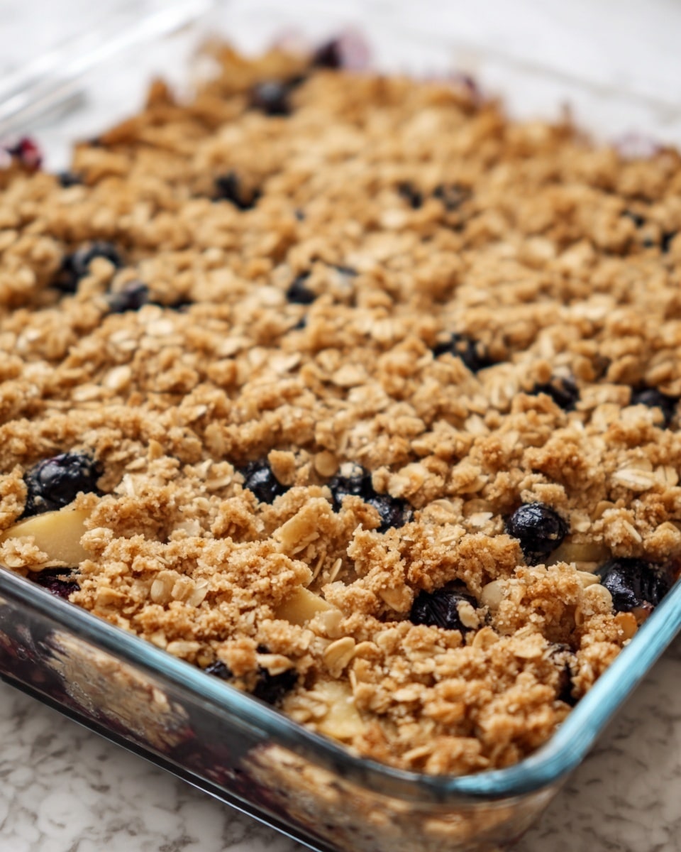 A close-up view of a square glass baking dish filled with a golden brown oat crumble on top. The crumble has a rough texture with visible pieces of oats and small chunks of light-colored fruit, along with dark blueberries scattered throughout. The surface looks slightly crispy and uneven, showing the baked oats covering the fruit layer underneath. The dish is placed on a white marbled texture. photo taken with an iphone --ar 4:5 --v 7