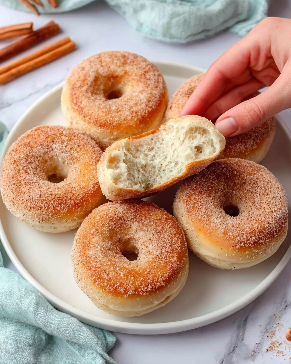 The image shows a white plate with six round doughnuts that have a rough sugar coating on top. The doughnuts are light golden brown on top and creamy beige on the sides, with a center hole in each one. A woman's hand is holding one doughnut broken in half, showing the soft, fluffy, white inside texture. The plate is on a white marbled surface with a light blue cloth partially visible on one side and some cinnamon sticks nearby. Photo taken with an iphone --ar 4:5 --v 7