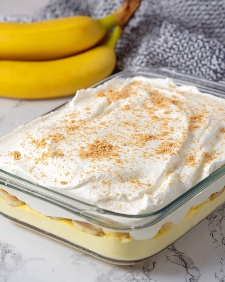 A rectangular glass dish holds a layered dessert placed on a white marbled surface. The bottom layer is pale yellow and smooth, topped with a thick, fluffy white layer that looks like whipped cream. The white top is sprinkled evenly with fine, light brown crumbs. In the background, a bunch of yellow bananas and a grey patterned cloth are partially visible. Photo taken with an iphone --ar 4:5 --v 7
