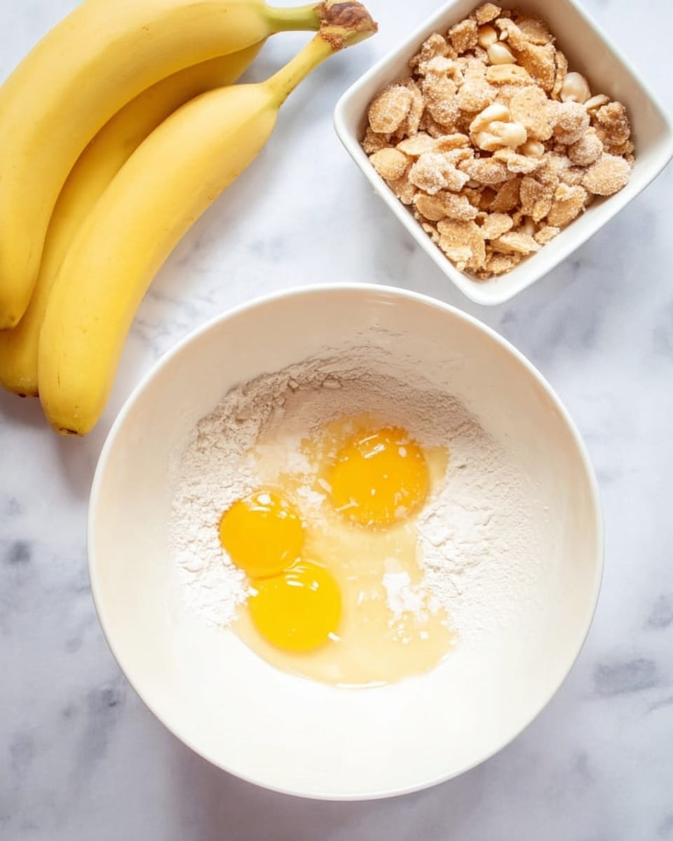A white mixing bowl on a white marbled surface contains three cracked raw eggs with bright yellow yolks sitting on top of white flour and a light brown liquid, creating a layered mix inside the bowl. To the right, there is a white square bowl filled with crushed beige and light brown cookies or nuts. To the left, three yellow bananas rest on the white marbled background. The overall scene is bright and clean with soft, natural light. Photo taken with an iphone --ar 4:5 --v 7