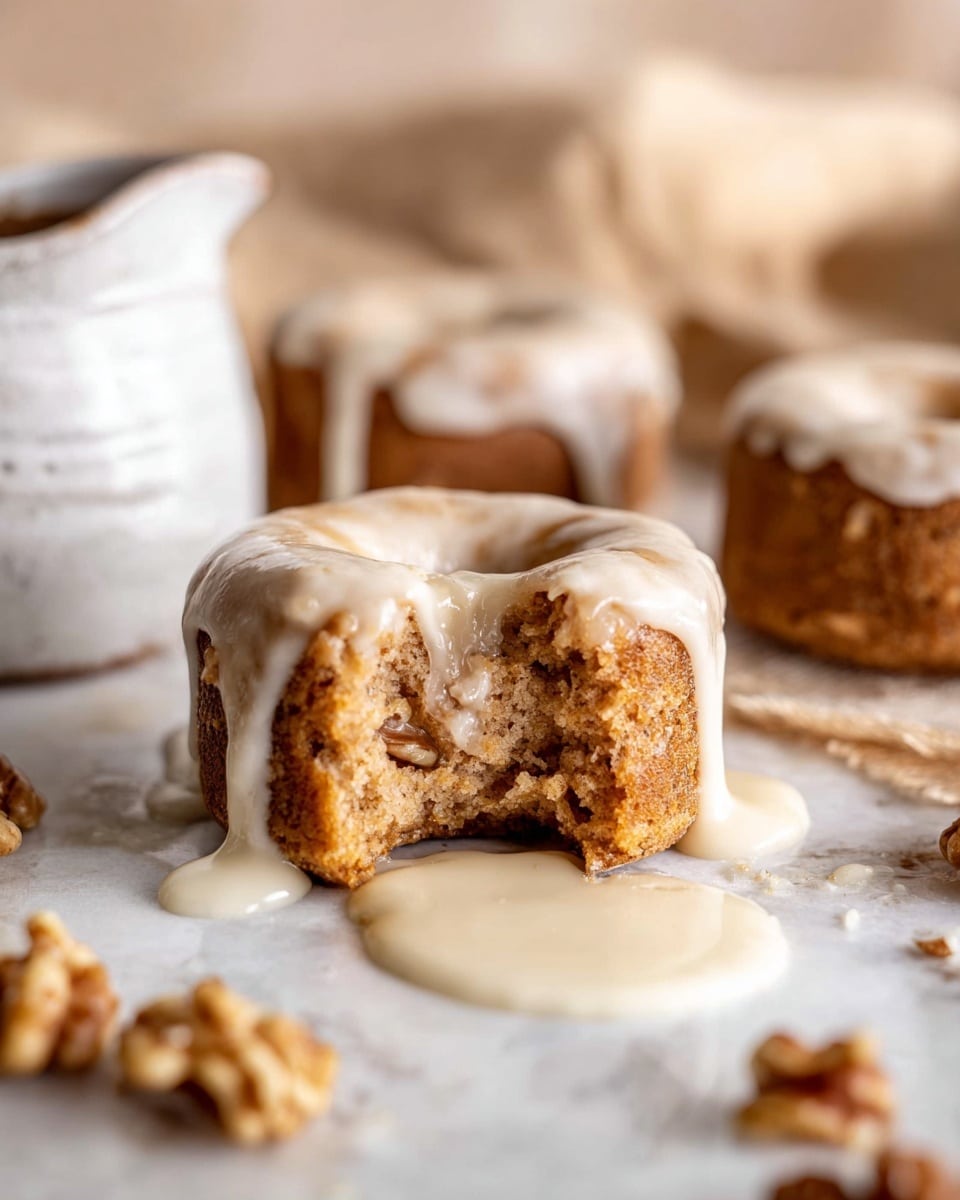A close-up view of a single small round cake with a light brown color and visible nut pieces inside, topped with a thick, creamy white glaze that drips down the sides and pools onto a white marbled surface. The cake has a bite taken out of it, showing its soft and crumbly texture inside. In the background, there are more small cakes with the same glaze, and pieces of walnuts scattered around the surface. A portion of a white ceramic jug with glaze drips is visible on the left side. The setting is softly lit and cozy, creating a warm and inviting feeling. Photo taken with an iphone --ar 4:5 --v 7