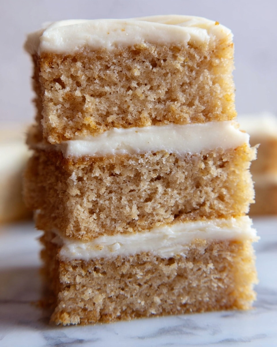The image shows a close-up of a stack of three thick square cake slices with a soft, crumbly texture and light brown color. Each slice is separated by a layer of smooth, creamy white frosting, with an additional thick layer of frosting on top of the stack. The background features a white marbled surface that contrasts with the cake’s warm tones. The focus is on the cake’s texture and frosting layers, showing fine details of the crumb and smoothness in the frosting. photo taken with an iphone --ar 4:5 --v 7