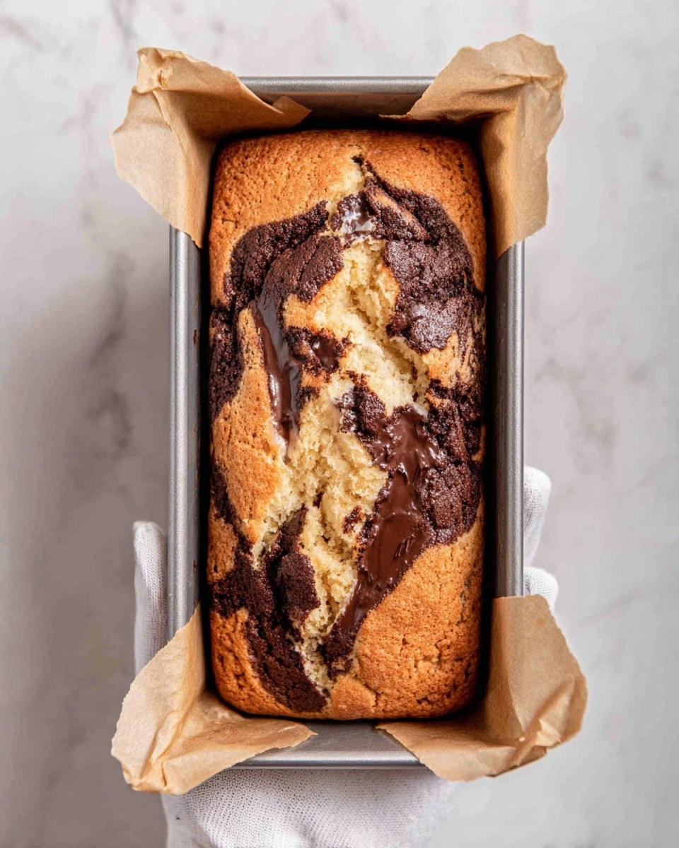 A rectangular loaf cake with a marbled pattern of two main layers, one light beige and one dark brown, swirled together across the top, creating an irregular patchwork of cracked and soft textures. The crust is slightly golden with a soft crumb visible where it has split at the center. The cake is inside a metal loaf pan with parchment paper lining the sides. A woman's hand wearing a white glove holds the pan from below against a white marbled surface. Photo taken with an iphone --ar 4:5 --v 7