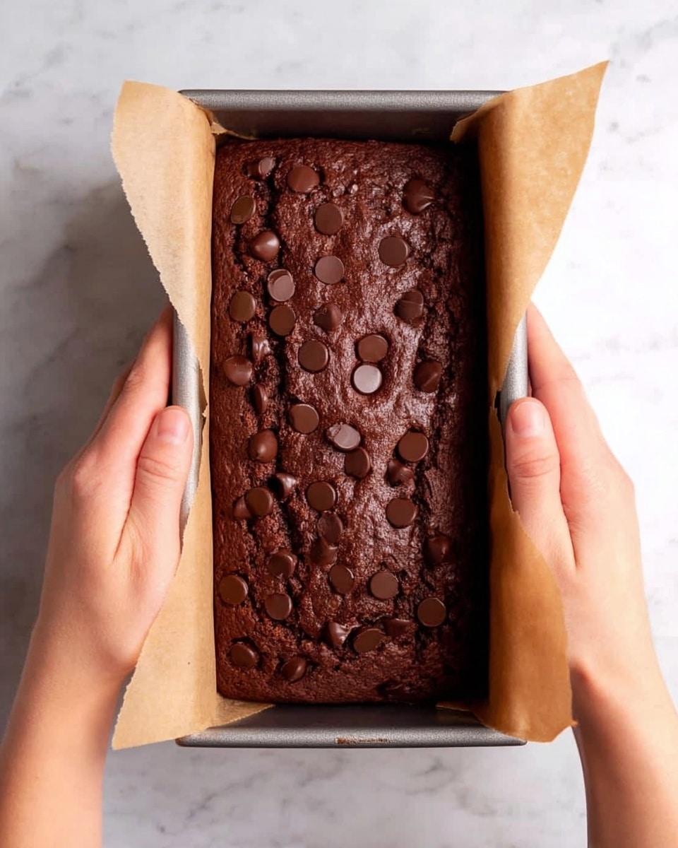 A rectangular chocolate cake loaf in a metal baking pan lined with parchment paper is shown being lifted by two woman's hands from each side. The cake has a rich dark brown color with a cracked top and is sprinkled with small round chocolate chips spread evenly across the surface. The background is a white marbled texture. Photo taken with an iphone --ar 4:5 --v 7
