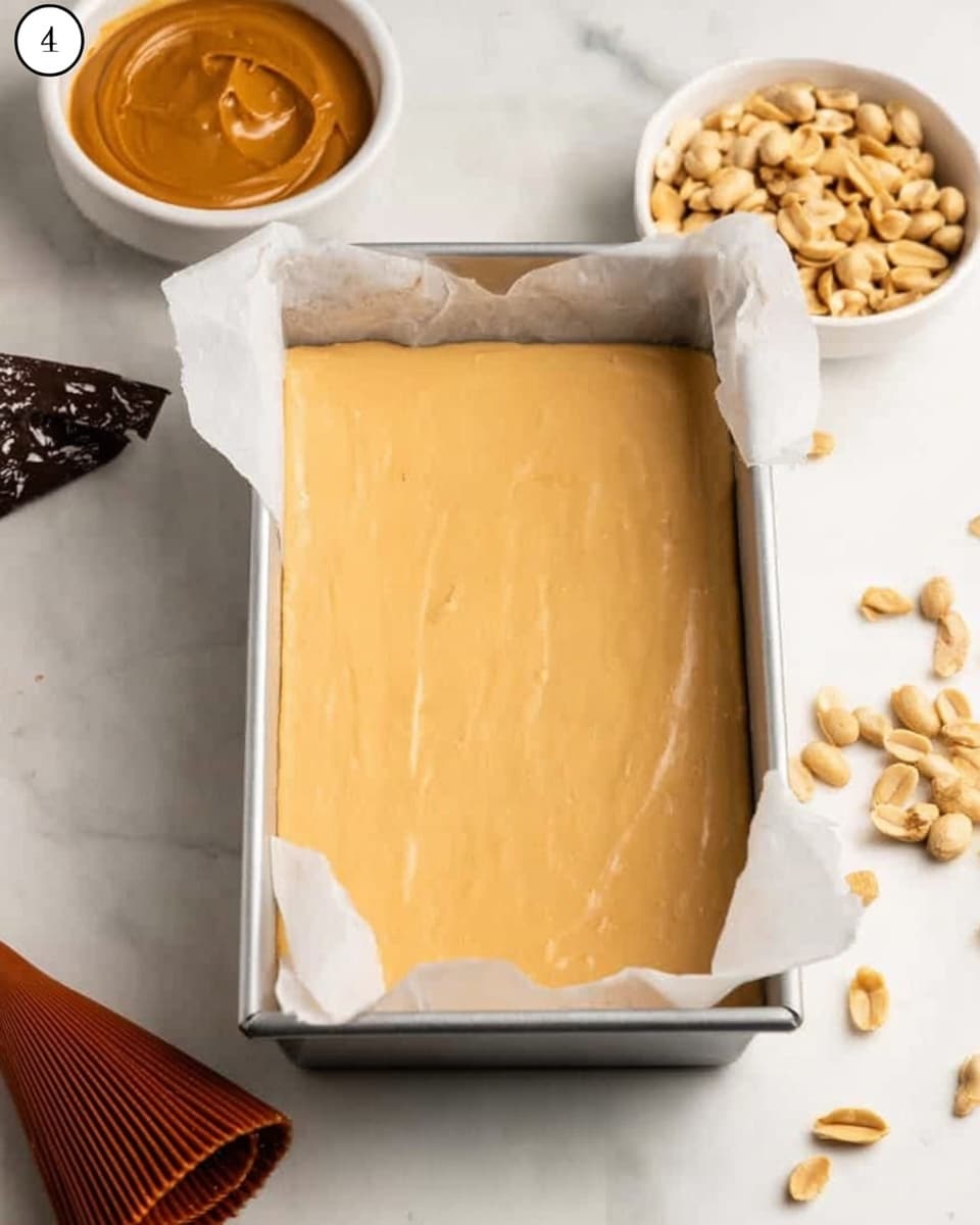 A rectangular metal baking pan lined with parchment paper is filled with a smooth, light tan batter, evenly spread to the edges. On the white marbled surface around the pan, there are clusters of shelled peanuts, a small white bowl filled with peanut butter, and a partially visible brown piping bag with a textured tip. The scene gives a simple, clean look focused on the creamy batter preparing to be baked. Photo taken with an iphone --ar 4:5 --v 7