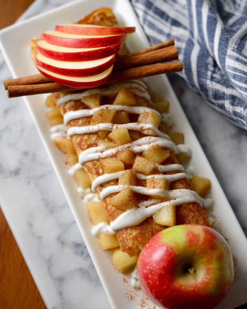 A long white rectangular plate holds a stack of small golden pancakes layered with cooked, diced apples that have a light brown, cinnamon dusting on top. White cream is drizzled across the stack in thin, uneven lines. At the top of the stack is a thin slice of red apple with a green stem, leaning against two cinnamon sticks placed diagonally. Next to the stack, a whole red apple with red and green patches rests on the plate. The plate is on a white marbled surface with a blue and white striped cloth partially visible on the side. photo taken with an iphone --ar 4:5 --v 7