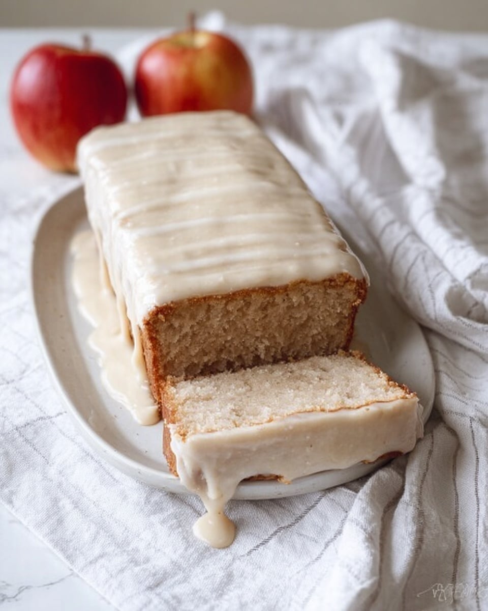 A rectangular loaf cake with a thick layer of creamy white icing covering the top and sides, with some icing dripping down onto the white plate beneath. The cake inside is light brown with a soft texture, shown by one slice cut at the front revealing its dense crumb. Two red apples sit in the background on the white marbled surface, next to a white cloth with light gray stripes. The lighting is soft and natural, giving a warm and cozy feel. photo taken with an iphone --ar 4:5 --v 7