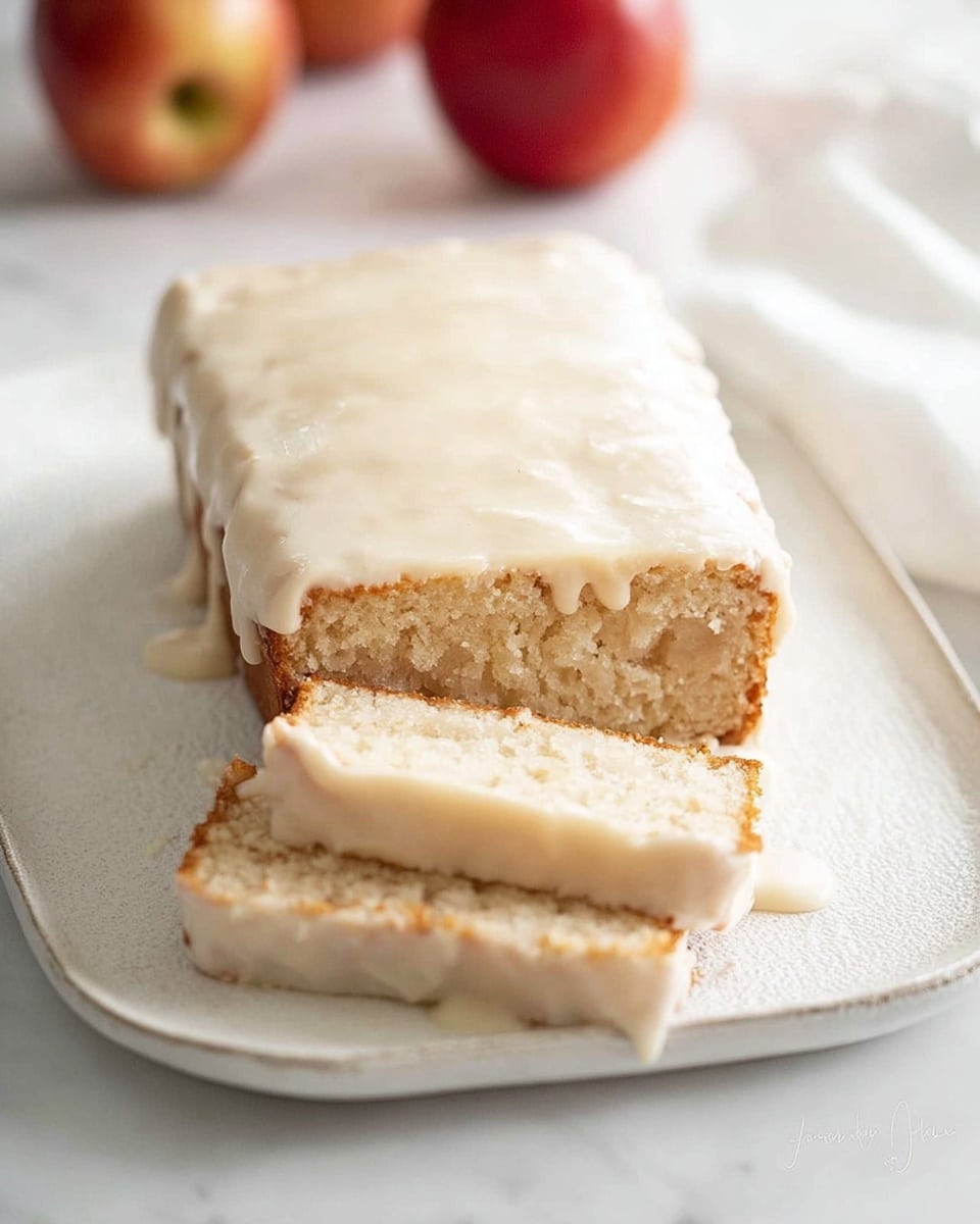 A rectangular loaf cake sits on a white, slightly textured plate with a thick, creamy white icing that covers the entire top and drips down the sides in smooth layers. Two slices have been cut from one end, revealing a dense, light beige crumb with small soft chunks inside. The icing on the sliced pieces is slightly uneven but thick, with a creamy texture that looks soft and moist. The background is a white marbled surface with two red apples blurred in the distance, adding color contrast. photo taken with an iphone --ar 4:5 --v 7
